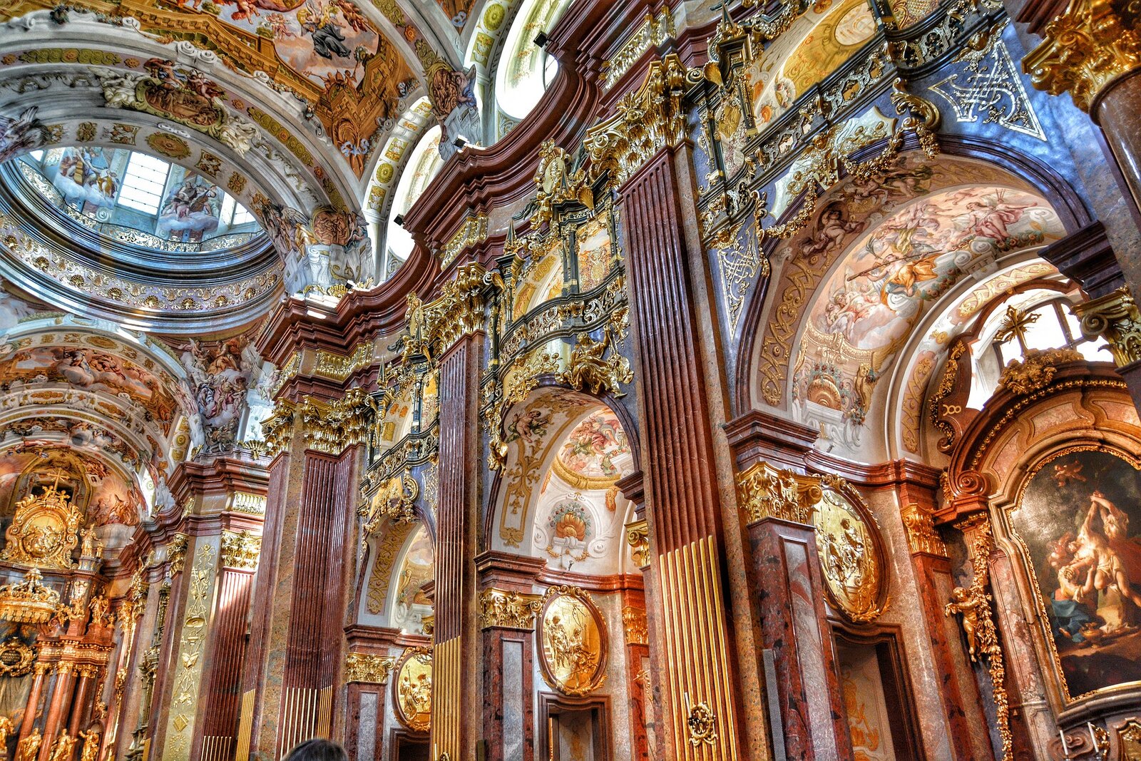 Ilustracja przedstawia wnętrze barokowego kościoła. Znajdują się duże kolumny, których głowice są złote. Na sklepieniach znajdują się kolorowe freski. Przy obrazach ze złotymi ramami i nad łukami są umieszczone złote rzeźby.