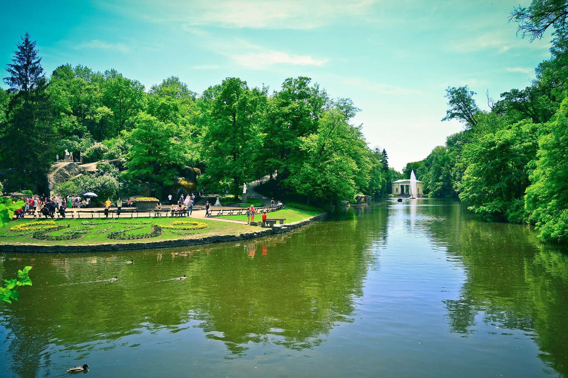 Ilustracja przedstawia zdjęcie fragmentu parku. Na pierwszym planie znajduje się akwen, w którym pływają kaczki. W tle po prawej stronie znajduje się niewielki pawilon, przed którym umieszczono fontannę.  Po lewej stronie znajduje się alejka parkowa z ławkami i klombami. Spacerują tam ludzie. W tle rosną wysokie zielone drzewa. Nad całym zdjęciem góruje niebieskie niebo.