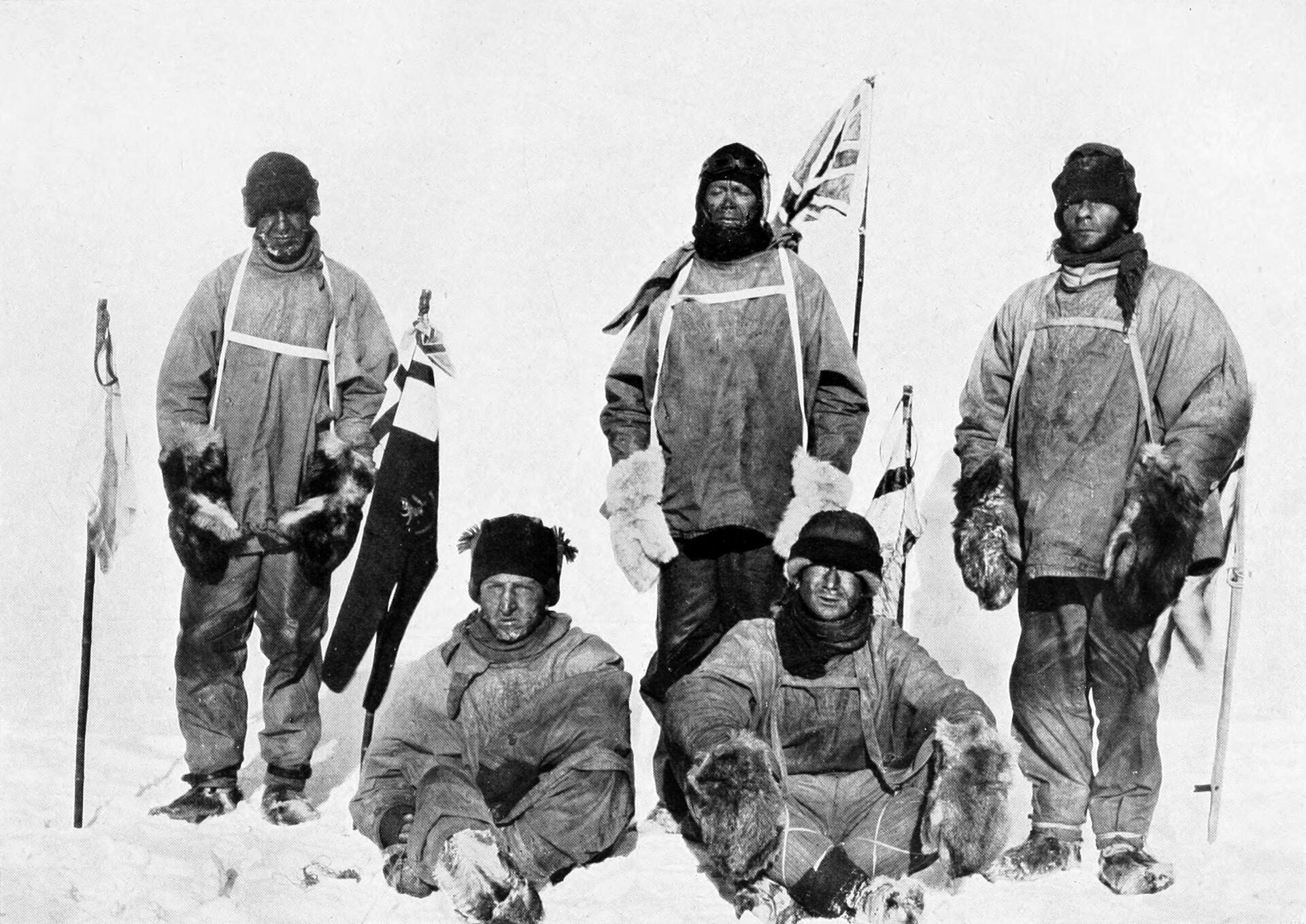 Na czarno‑białym zdjęciu pięciu mężczyzn w ciepłych strojach i futrzanych rękawicach, z ogorzałymi twarzami. Za nimi wbita w śnieg flaga brytyjska.