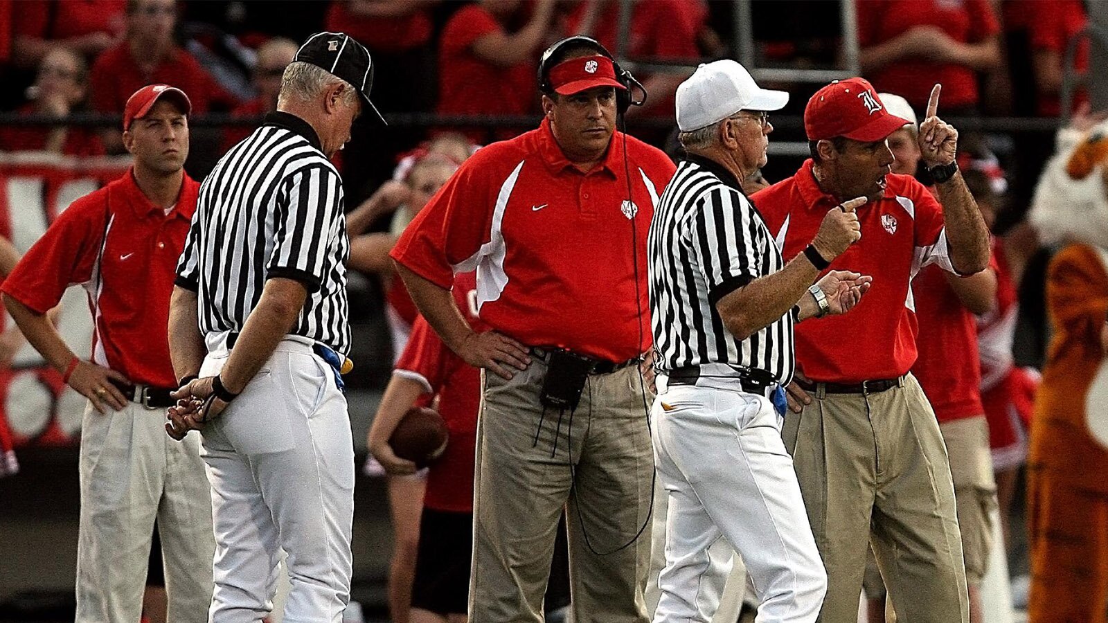 Zdjęcie przedstawia grupę stojących mężczyzn. Trzech z nich ma koszulki w kolorze czerwonym, a dwóch – koszulki w czarne i białe paski. Wszyscy mają czapki z daszkiem na głowie. Mężczyzna stojący pośrodku ma założone na głowę słuchawki z mikrofonem. Mężczyźni gestykulują.