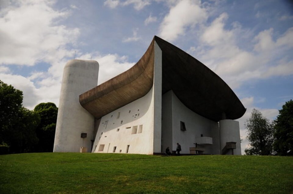 Zdjęcie przedstawia katedrę Notre Dame w Ronchamp, we Francji, zaprojektowaną przez Le Corbusiera. Ma masywną bryłę, na planie trójkąta, z niewielkimi okienkami. Ciekawą formę ma dach, przypominający miękkie nakrycie głowy, zbiegające się z przodu ostrym kantem.