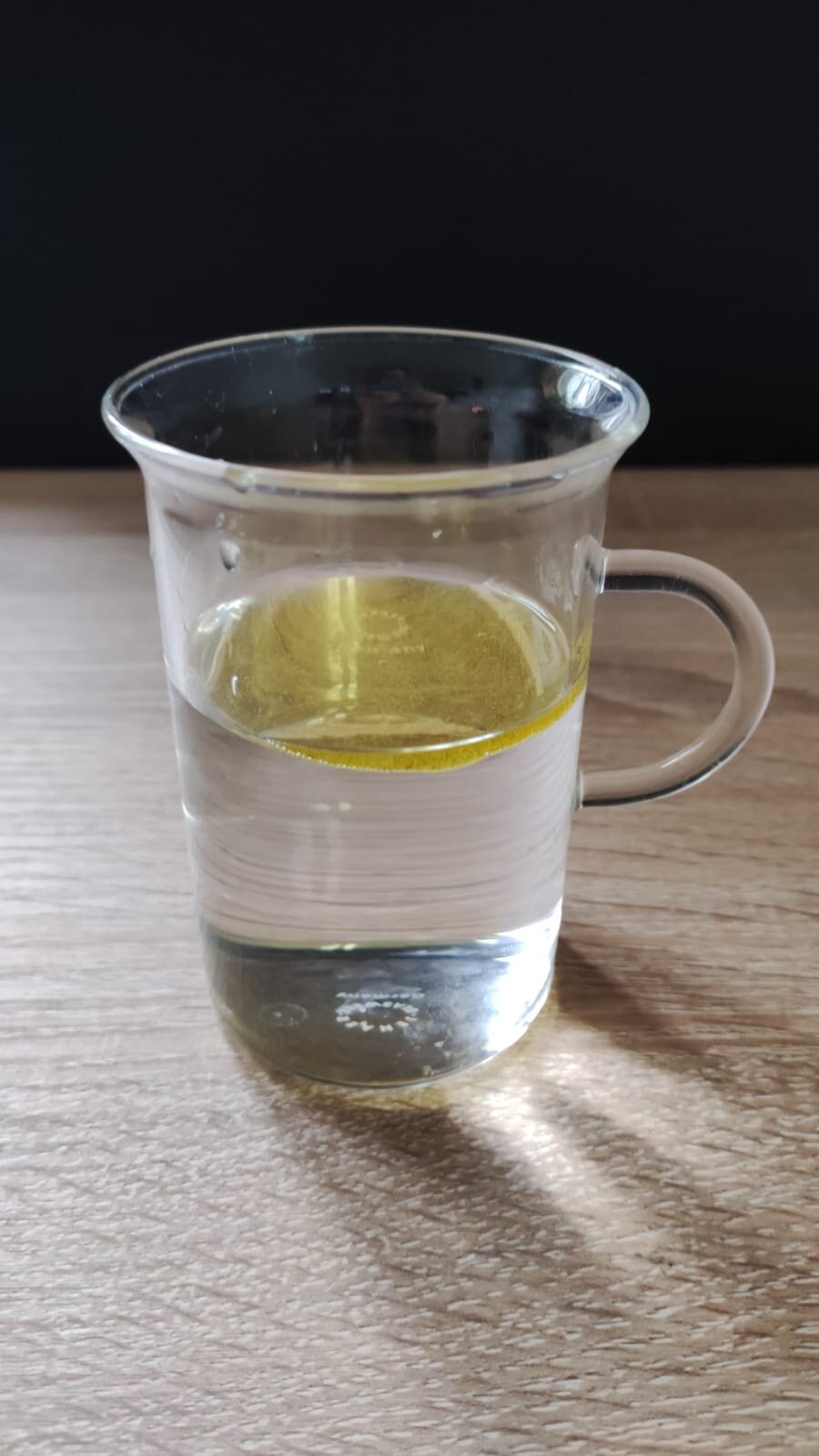 Na fotografii znajduje się szklanka wypełniona do połowy wodą. U góry unosi się plama żółtego oleju, tworząc warstwę na powierzchni wody.