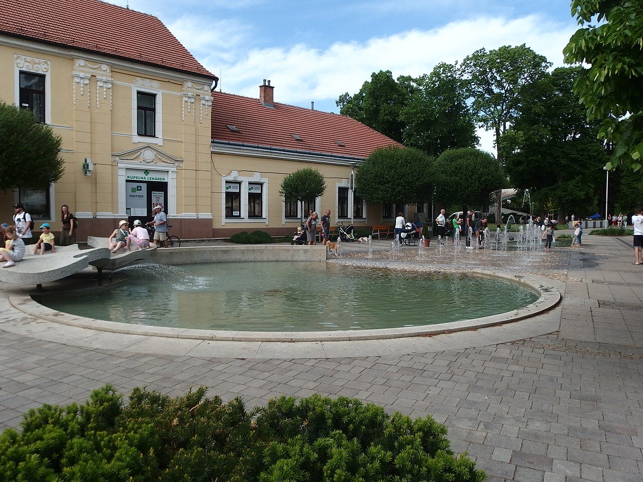 Na zdjęciu na pierwszym planie oczko wodne i fontanna w mieście. Wokół dzieci chlapią się wodą. Za obiektem znajduje się zółty budynek z czerwonym dachem. Na nim znajdują się białe ozdoby. Obok budynku i dookoła placu na którym znajduje się fontanna rosną drzewa. Po placu przechadzają się ludzie. 