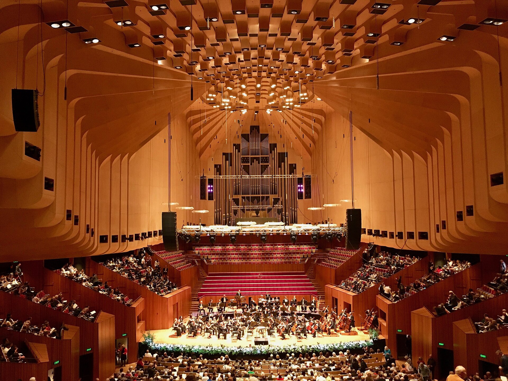 Zdjęcie przedstawia sale koncertową Sydney Opera House w czasie koncertu. Na środku zdjęcia przedstawiona jest scena muzyczna, na której znajduje się zespół opery. Wokół sceny znajduje się widownia. Widownia znajduje się na różnych poziomach sali. Sala koncertowa jest koloru brązowego oraz czerwonego.  