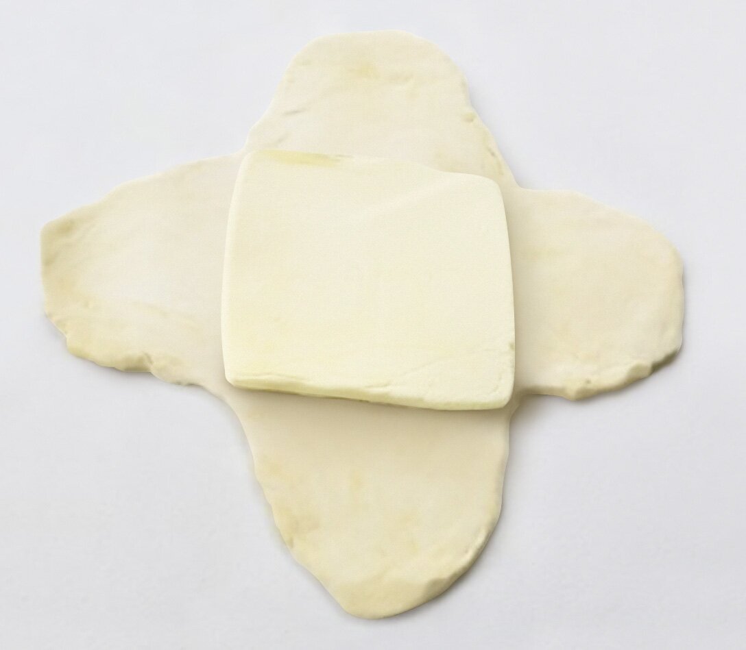 Ciasto rozwałkowane w kształt czteroramiennej gwiazdy, na środku znajduje się położona, kwadratowa kostka ciasta.