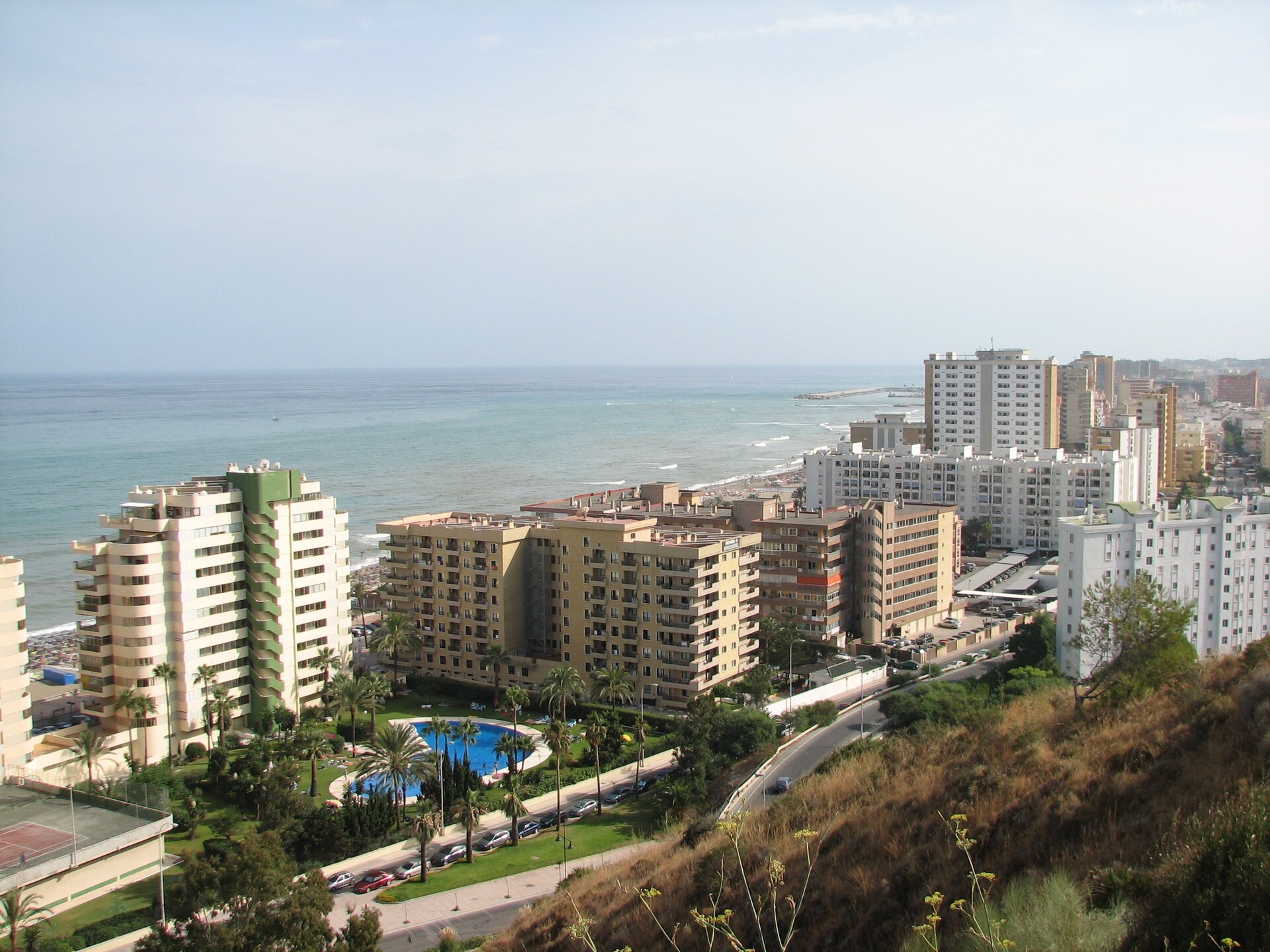 Na zdjęciu wysokie bloki przy brzegu morza. Pomiędzy budynkami bogata roślinność, basen.