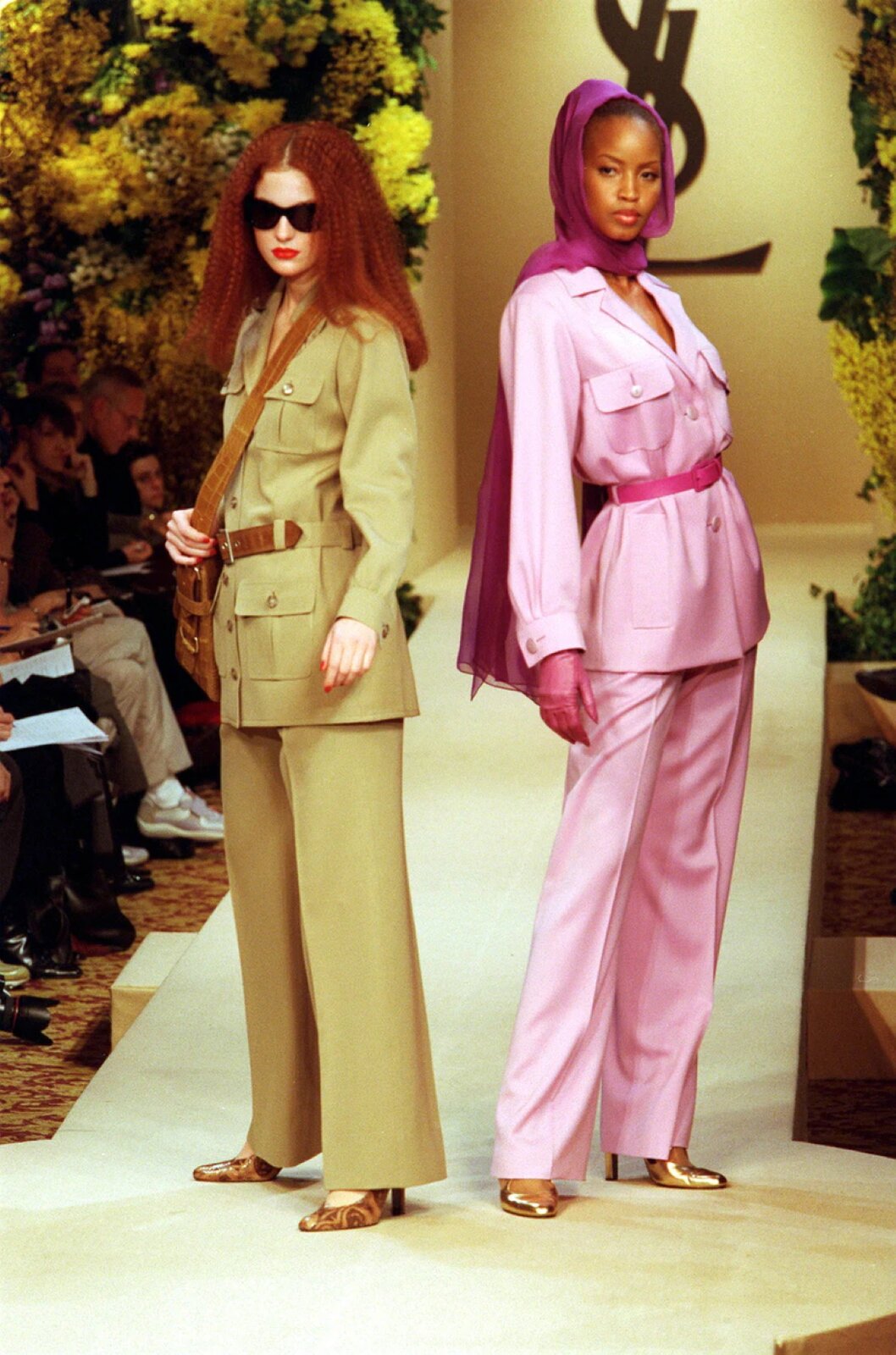 Fotografia przedstawia dwie modelki stojące obok siebie na wybiegu. Obie mają na sobie obszerne marynarki z paskiem w talii oraz eleganckie, rozszerzane ku dołowi spodnie i buty na obcasach. Komplet rudowłosej kobiety stojącej z lewej strony jest jasnobrązowy, ma ona na sobie również ciemne okulary. Druga, ciemnoskóra kobieta ma włosy owinięte fioletową chustą, a jej ubrania są różowe. W tle widać siedzących ludzi oraz kwiatowe dekoracje pokazu mody.