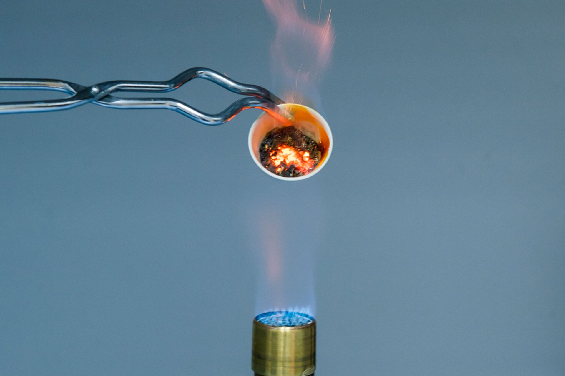 Zdjęcie przedstawia gazowy palnik laboratoryjny, nad którym znajduje się końcówka poziomo ustawionych szczypiec trzymających mały tygielek ogrzewany w płomieniu i obrócony wlotem w kierunku aparatu. W tygielku znajduje się rozżarzona mieszanina siarki z żelazem.