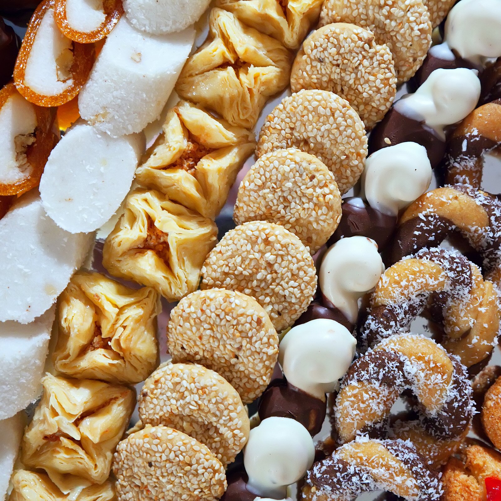 Zdjęcie przedstawia ułożone w rzędach różnego rodzaju ciastka. Widoczne są kawałki baklawy, okrągłe makaroniki obsypane sezamem i małe pączki z dziurką. 