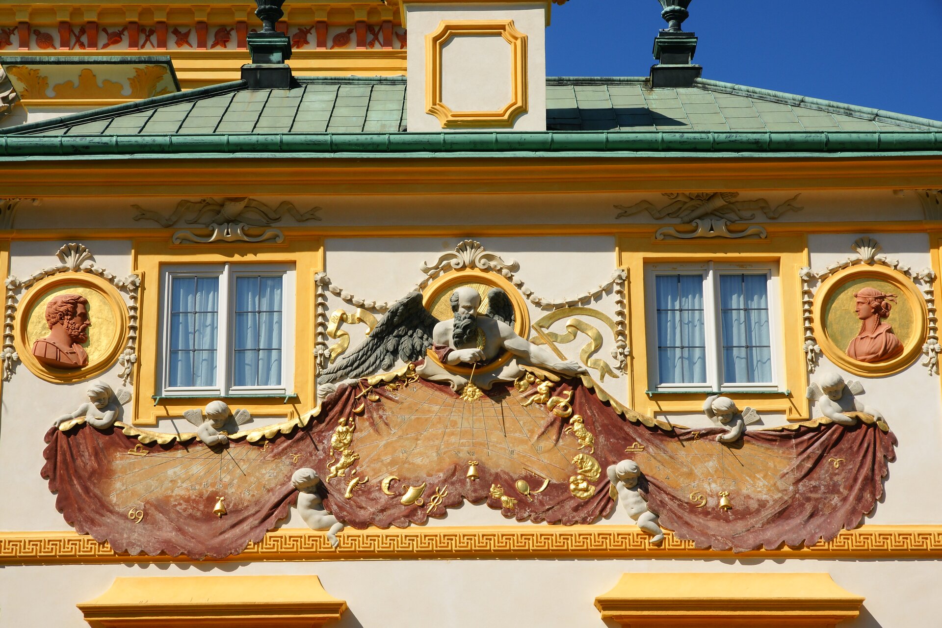 Fotografia autorstwa Bosia2011 przedstawia widok na zegar słoneczny, który znajduje się na elewacji Pałacu Jana III Sobieskiego. Zegar składa się z kilku części – Figury mężczyzny w podeszłym wieku, który ma bardzo długą brodę i siwe skrzydła i spogląda się na kupidyny. Poniżej figury znajduje się złota część płaskorzeźby imitująca płótno na którym znajdują się wskazówki zegara słonecznego. 