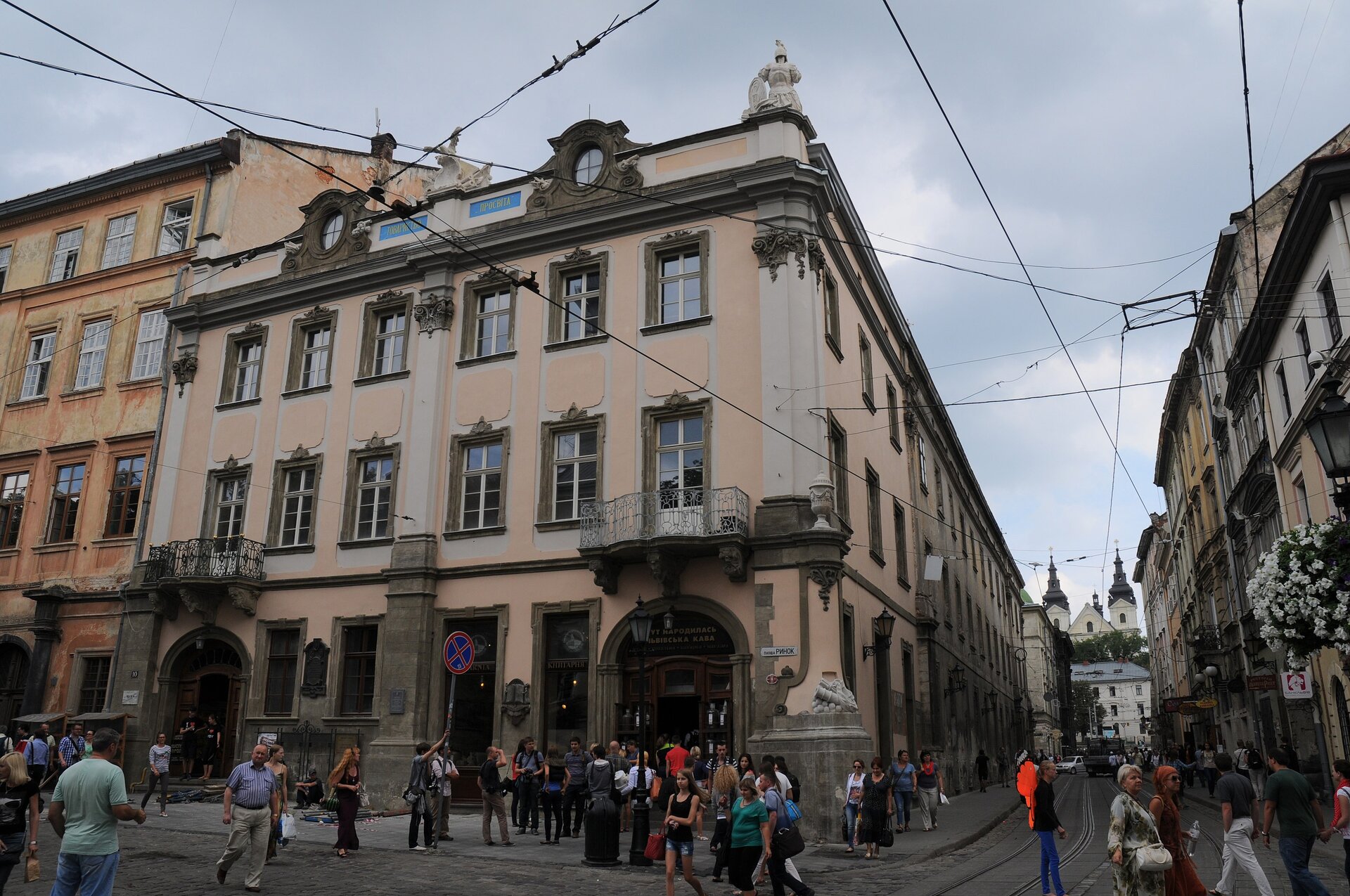 Ilustracja przedstawia pałac Lubomirskich we Lwowie, widok współczesny. Budynek powstał w miejscu dwóch kamienic i pięciu domów. Jest on trzykondygnacyjny. Ma brązowe okucia i zdobienia, elewacja jest koloru beżowego. Ma niewielkie okna, figury na szczycie.  