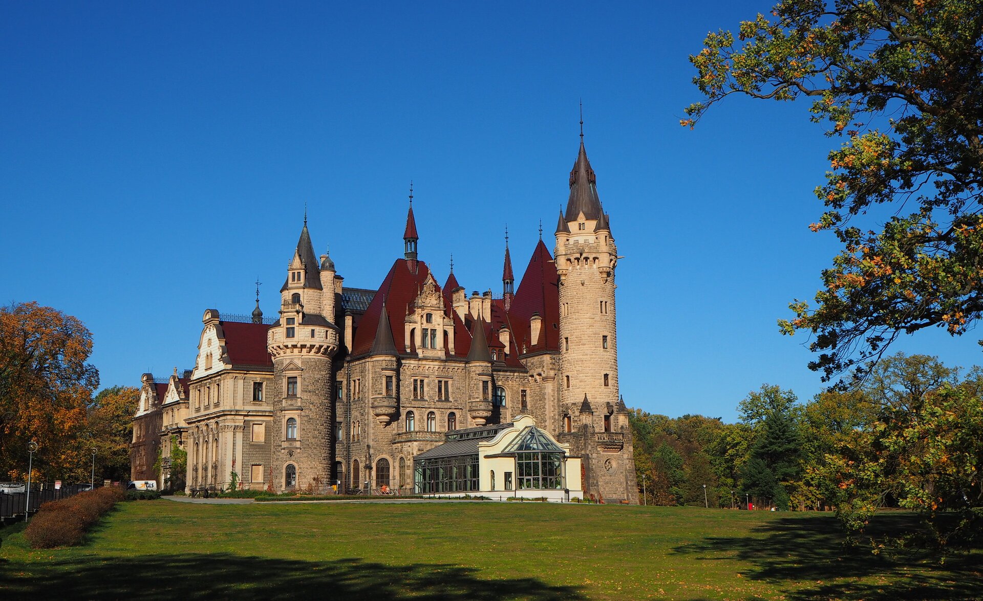 Zdjęcie przedstawia pałac w Mosznej. Budowla składa się z różnych, przylegających do siebie brył geometrycznych. Są to części budynku w kształcie prostopadłościanów i wieże w kształcie walców. Mury są wykonane z kamiennych cegieł o nieregularnym kształcie, w kolorze szarobrązowym. Pałac ma wiele małych okienek. Zwieńczony jest strzelistymi dachami w kolorze ciemnoczerwonym i brązowym. Z przodu znajduje się także niskie, oszklone pomieszczenie, w którym rosną rośliny. Dokoła pałacu rośnie przystrzyżona trawa oraz drzewa.