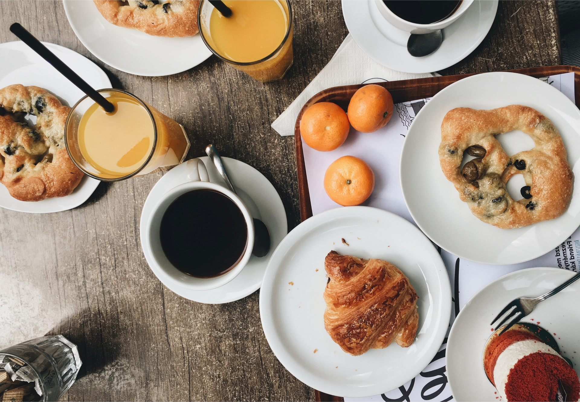 Zdjęcie, zrobione z góry, przedstawia stół, na którym ustawione są naczynia i produkty śniadaniowe. W szklankach ze słomką jest sok pomarańczowy, w filiżankach czarna kawa. Są też talerze, na dwóch z nich leżą precle z oliwkami, na kolejnym francuski rogalik i na ostatnim tiramisu. Na stole leżą też 3 mandarynki.