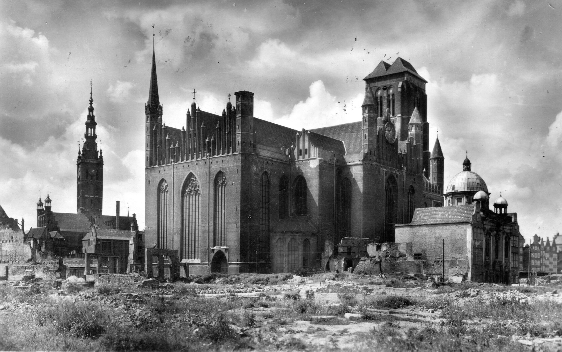 Zdjęcie przedstawia ul. Świętego Ducha w Gdańsku po zniszczeniach II wojny światowej. W centralnej części widoczne są budynki Kaplicy Królewskiej, kościoła Mariackiego i Ratusza Głównego Miasta.