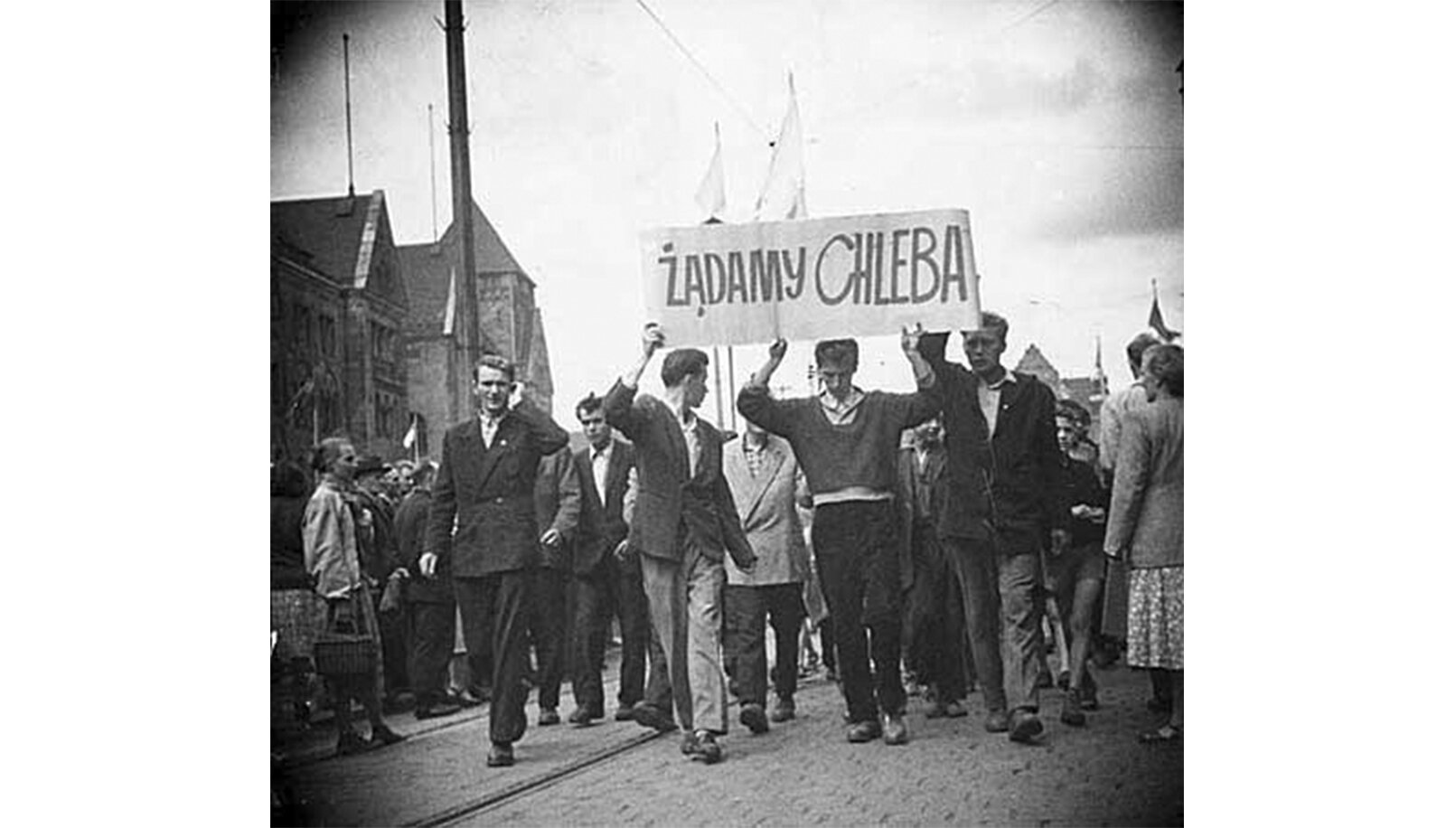 Czarno-biała fotografia przedstawiająca manifestację na ul. Armii Czerwonej (obecnie Święty Marcin) w Poznaniu w 1956 roku. Demonstranci niosą transparent z napisem: Żądamy chleba .