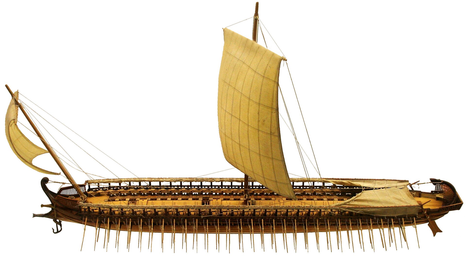 Fotografia przedstawia makietę starożytnego okrętu na białym tle. Długi i wąski okręt ma dwa żagle: jeden duży na środku i mniejszy na dziobie. Z boku okrętu widoczne są rzędy wioseł.  