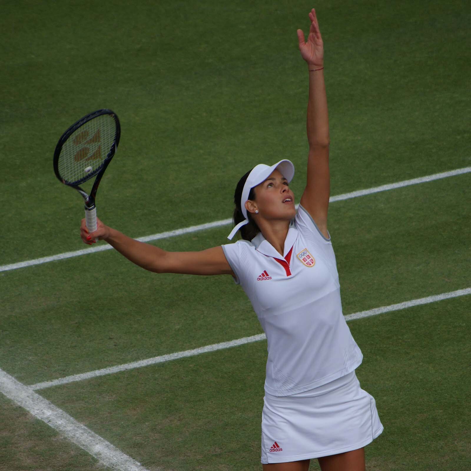 Zdjęcie przedstawia tenisistkę z rakieta uniesioną do serwu na korcie trawiastym.