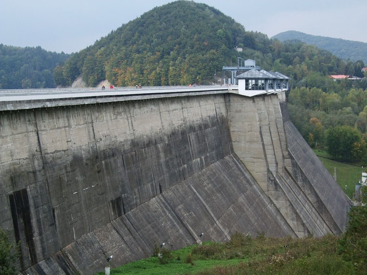 Zdjęcie przedstawia zaporę Zbiornika Solińskiego. Zapora jest wysoka i zbudowana z betonu. Na jej szczycie znajduje się budynek. W tle widoczny krajobraz górski. 