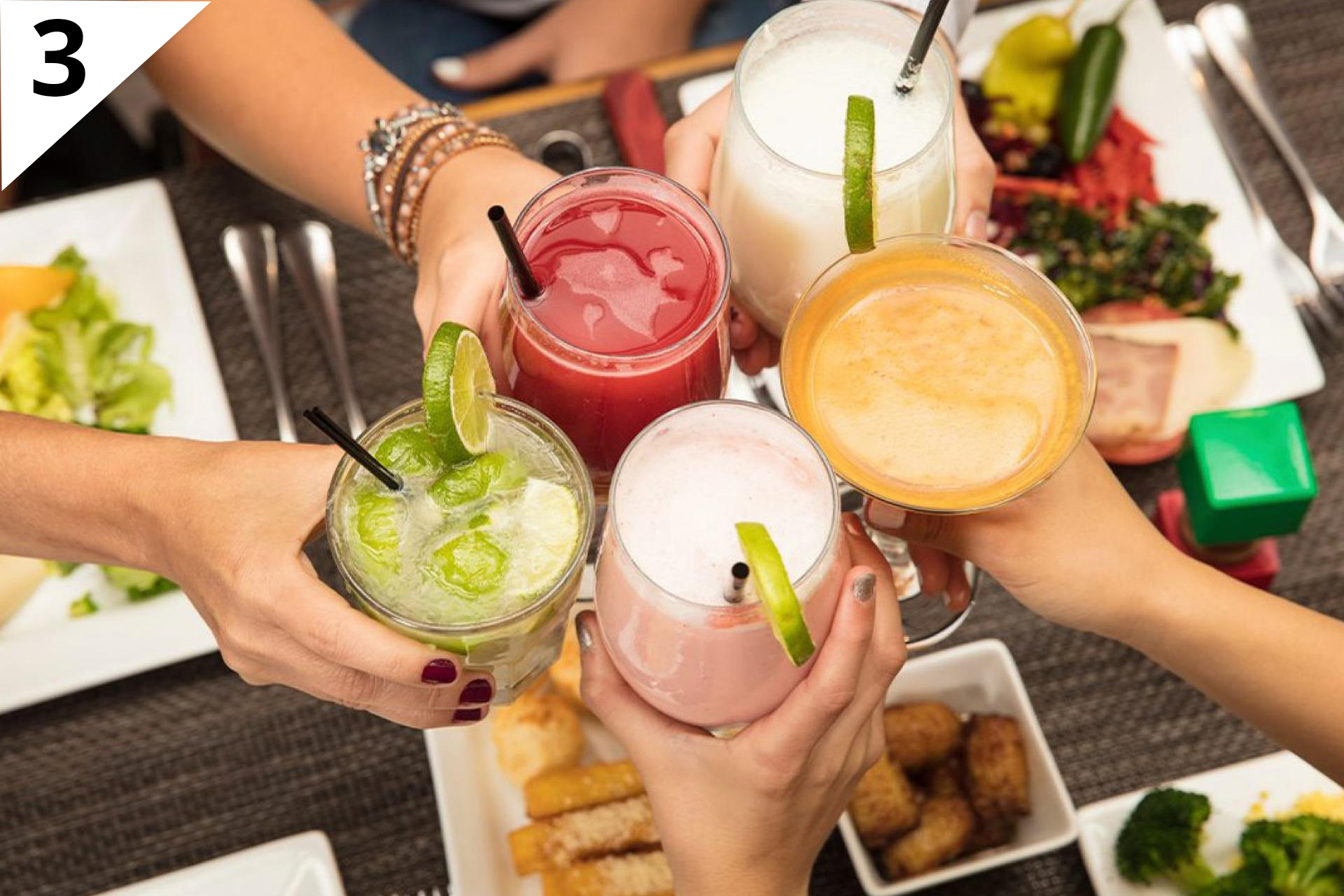 Zdjęcie przedstawia pięć różnych dłoni trzymających nad zastawionym stołem szklanki lub kieliszki z kolorowymi koktajlami owocowymi.