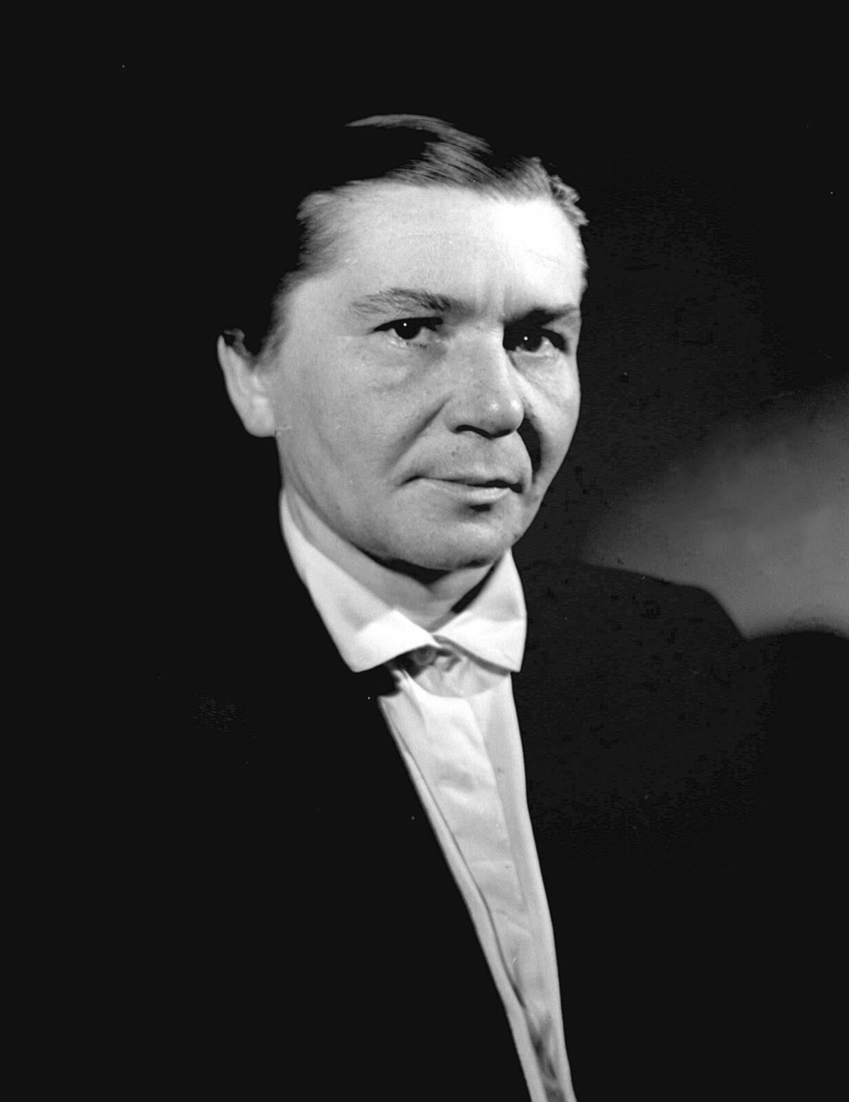 Na fotografii widoczna jest reżyser i scenarzystka – Wanda Jakubowska, która została uchwycona jako kobieta w średnim wieku o ciemnych włosach zaczesanych do tyłu. Ma neutralny wyraz twarzy i spogląda w obiektyw. Pani reżyser ubrana jest w ciemny żakiet oraz białą koszulę z kołnierzem. Na jej twarzy widoczne są zmarszczki. Tło jest ciemne. 