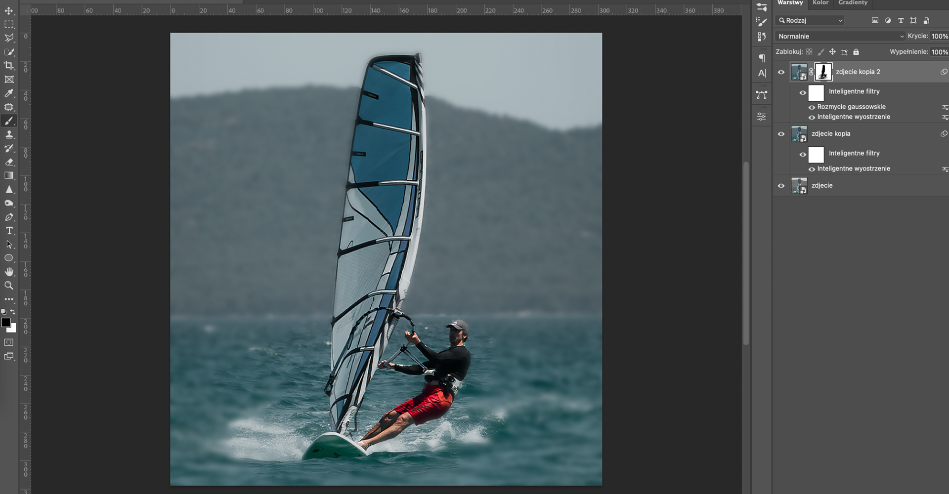 Ilustracja przedstawia okno programu. W obszarze roboczym jest zdjęcie mężczyzny na windsurfingu. Wyraźna jest postać mężczyzny, natomiast tło i woda są rozmyte. Woda jest wyraźna wyłącznie w bliskim sąsiedztwie windsurfingu i nieco  z tyłu. Po prawej stronie jest panel dotyczący warstw.  Tam zdjęcie kopia 2, zdjecie kopia, zdjecie. Zaznaczono zdjecie kopia 2.  