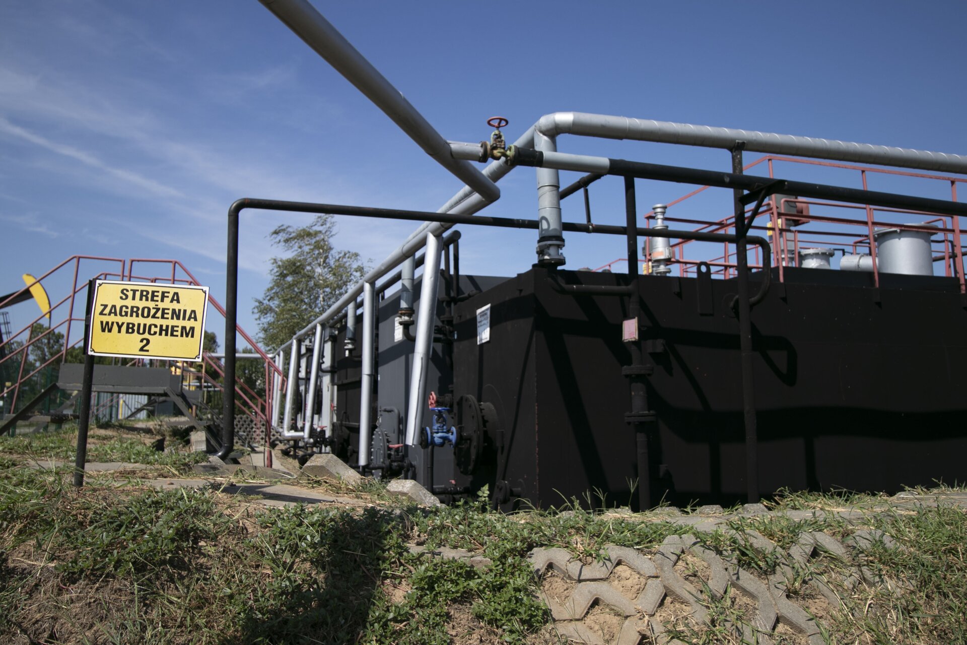Zdjęcie przedstawia zespół prostopadłościennych zbiorników magazynowych wraz z orurowaniem oraz oznaczeniem strefy zagrożenia wybuchem. Są one ze sobą połączone. Na górze każdego z nich zamontowana jest platforma z barierką, umożliwiająca poruszanie się pracownikom i dostęp do wnętrza zbiornika. Po lewej stronie znajduje się tabliczka. Na żółtym tle czarny napis. Strefa zagrożenia wybuchem dwa.