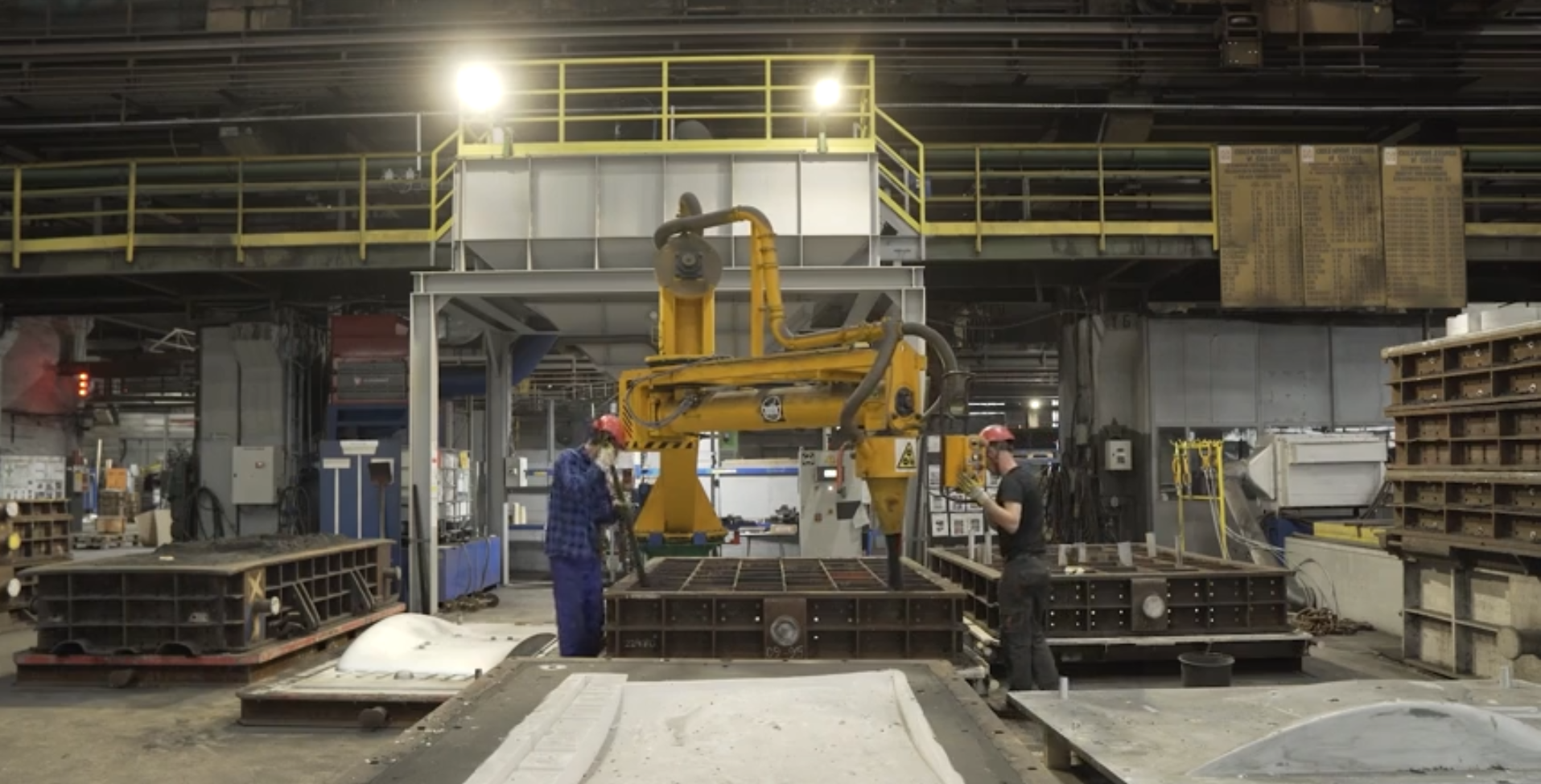 Na zdjęciu widoczna jest mieszarko‑nasypywarka podczas wypełniania. W centralnej części zdjęcia znajduje się żółte urządzenie. Urządzenie składa się z ruchomych ramion. Urządzenie jest obsługiwane przez dwóch pracowników. Pracownicy mają stroje roboty i czerwone kaski.Urządzenie znajduje się w dużej hali przemysłowej. Na górze hali widoczna jest galeria. Galeria jest zabezpieczona żółtą barierką.