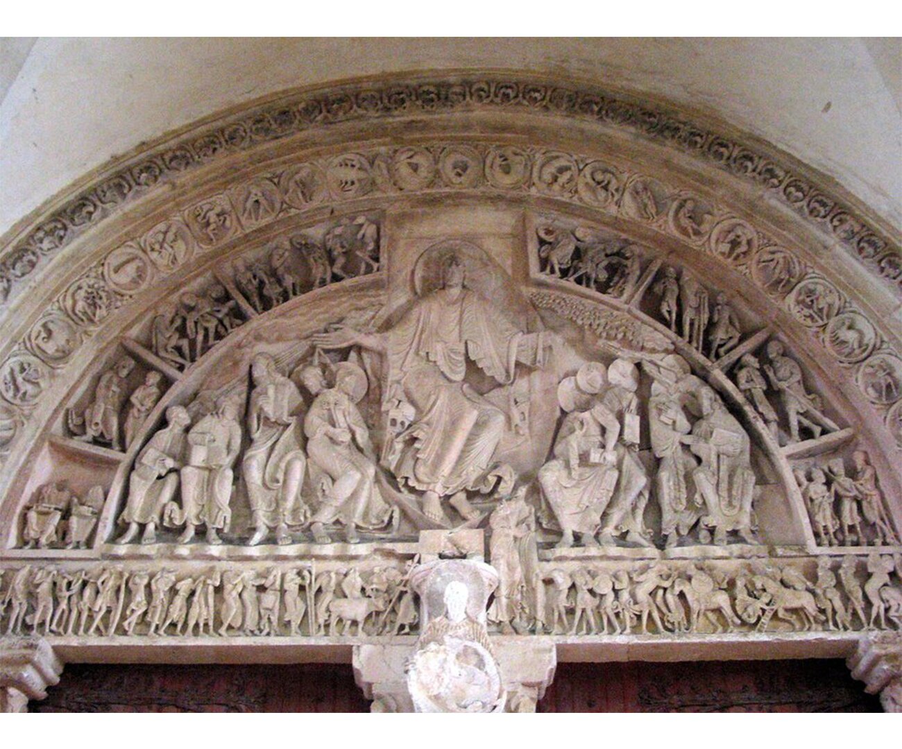 Fotografia barwna przedstawiająca Tympanon kościoła w Vazley. Uwidocznione zostały wyżłobienia sceny z główną postacią, największą w środku. W tle dużo wyżłobionych postaci.