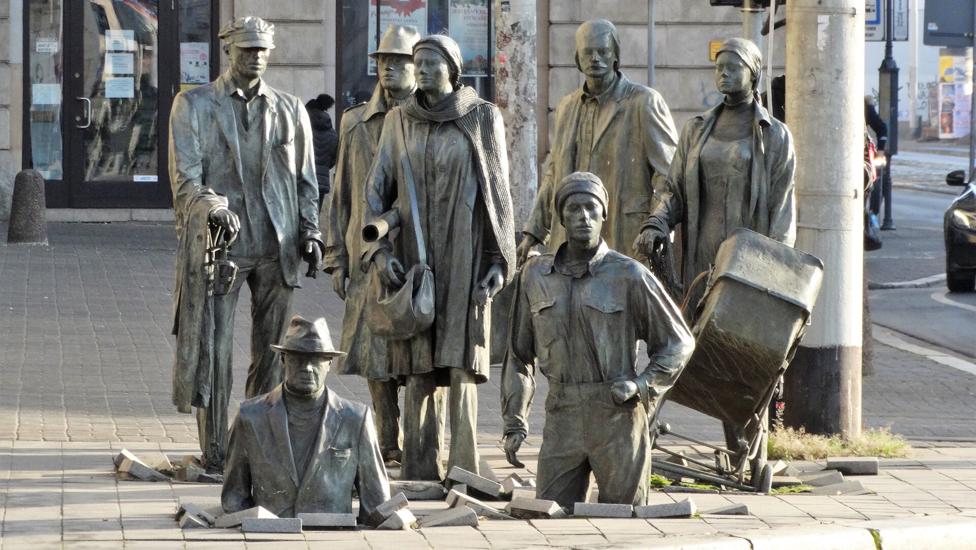 Zdjęcie przedstawia pomnik autorstwa Jerzego Kaliny, pod tytułem „Pomnik anonimowego przechodnia”. Dzieło przedstawia ludzi, postacie z brązu naturalnej wielkości. Na fotografii ukazanych jest siedem osób. Na pierwszym planie, na środku znajdują się dwaj mężczyźni, którzy znikają w chodnikowych płytach. To sprawia, że tworzy się iluzja zapadającej się ziemi.  Za mężczyznami  po prawej stronie grupy figur można wyróżnić kobietę z dziecięcym wózkiem oraz mężczyznę z walizką. W środku pomiędzy mężczyznami  znajduje się kobieta z torebką oraz zwojem papieru. Po lewej stronie za mężczyzną  znikającym w płytkach stoi starszy mężczyzna z laską, a tuż obok niego mężczyzna w kapeluszu. Cała kompozycja figur znajduje się na chodniku, tuż przy ulicy. W oddali widać budynki.  