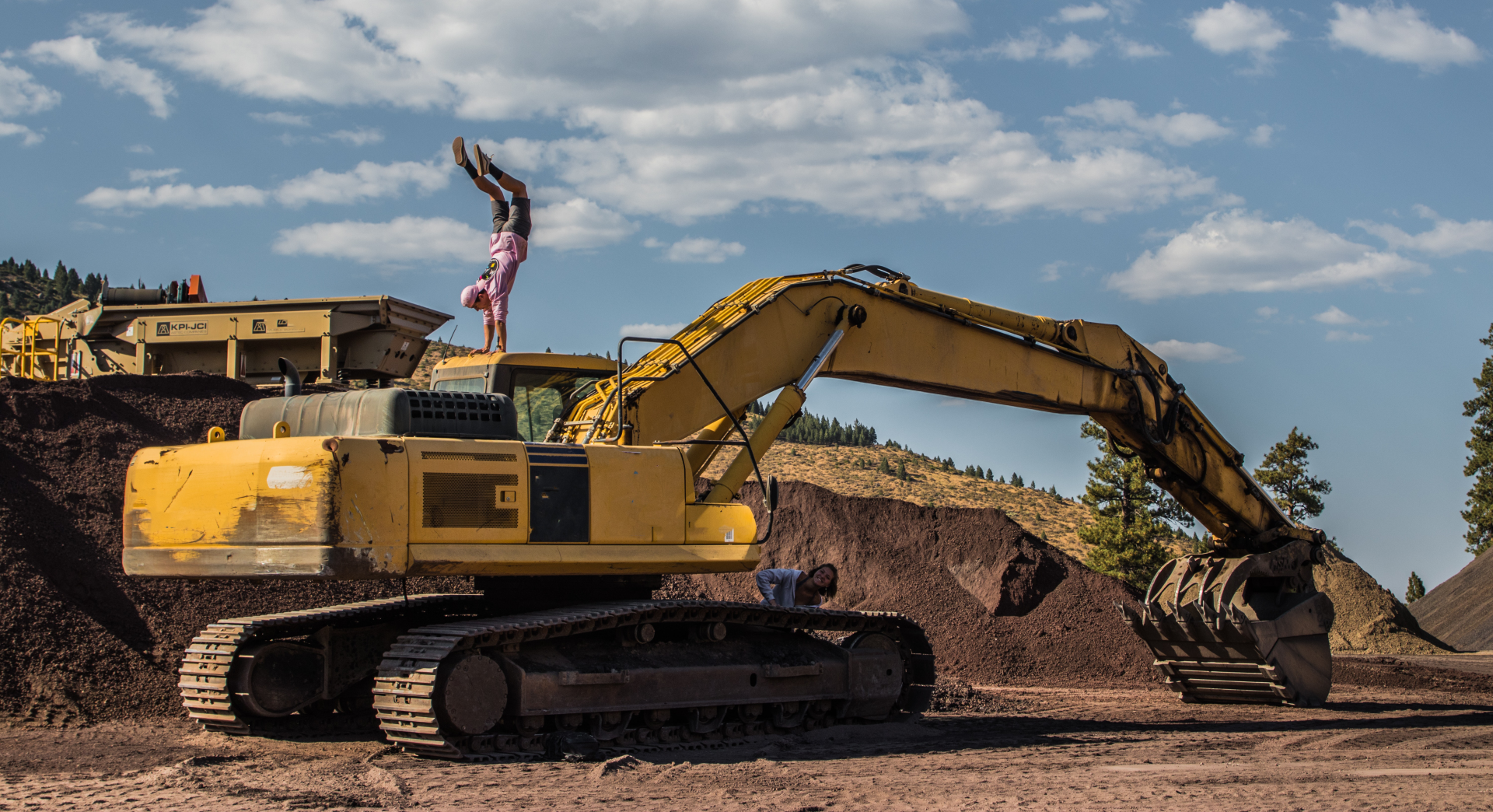 Fotografia przedstawia żółtą koparko‑ładowarkę na gąsienicach, na terenie budowy.