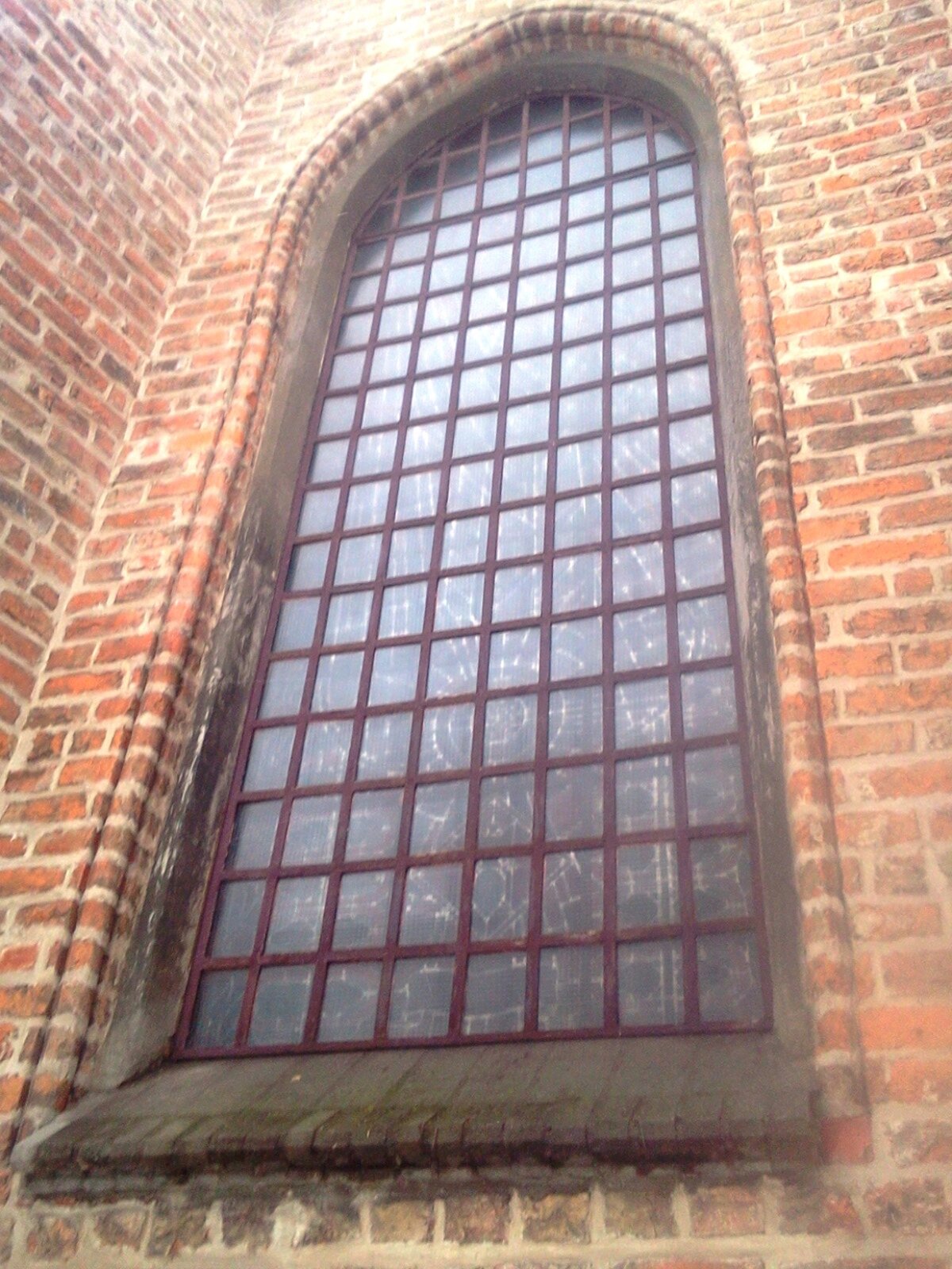 Fotografia przedstawia fragment budynku zbudowanego z pomarańczowych cegieł oraz zbliżenie na witraż od zewnątrz. Okno podzielone zostało na sekcje składającą się ze 134 elementów - każdy element to fragment witrażu. Okno posiada ukośny parapet. 