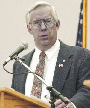 Zdjęcie przedstawia mężczyznę w średnim wieku. Stoi i przemawia z mównicy. Mężczyzna ma krótkie, siwe włosy z zaczesaną na bok grzywką. Nosi okulary. Ubrany jest w garnitur. Kolor skóry twarzy mężczyzny jest szary.