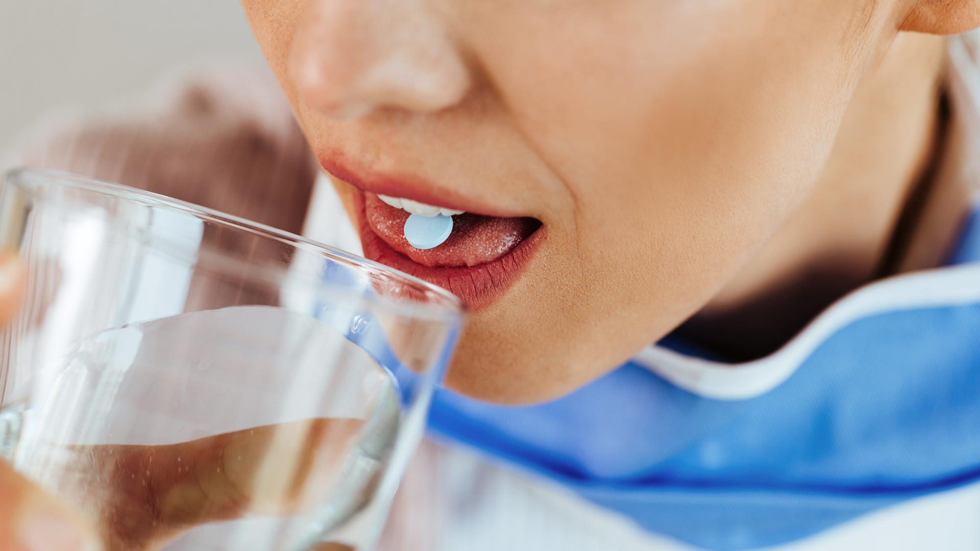 Zdjęcie przedstawia zbliżenie na usta osoby. W ustach znajduje się tabletka. Osoba trzyma szklankę z wodą.