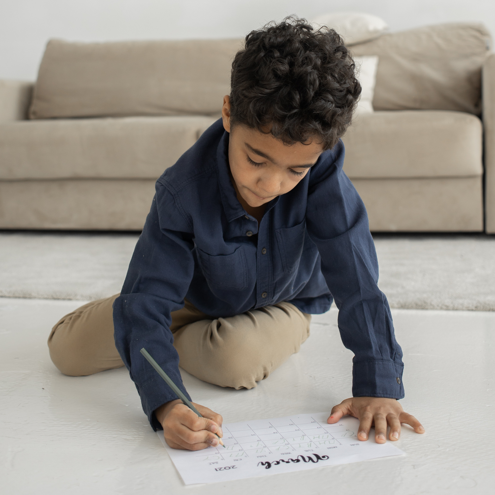 Zdjęcie przedstawia chłopca, w wieku około 11 lat, siedzącego na podłodze w pokoju z sofą. Chłopiec trzymając w prawej ręce szarą kredkę, nachyla się nad kartką na której jest napis "MARCH" a pod nim tabela ukazująca dni.