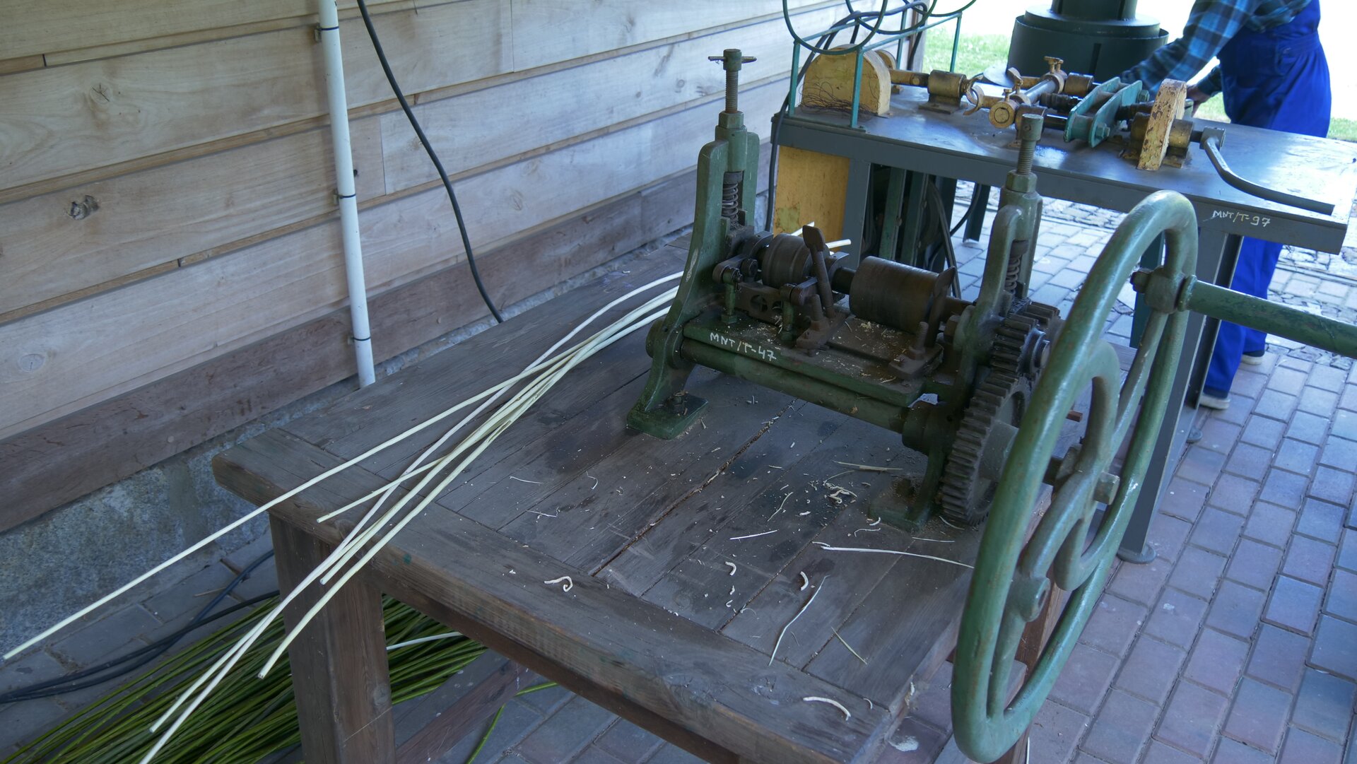 Zdjęcie przedstawia taśmiarkę ręczną stojąca na zewnątrz budynku. Urządzenie stoi na drewnianym stole i posiada duże koło zamachowe. 
