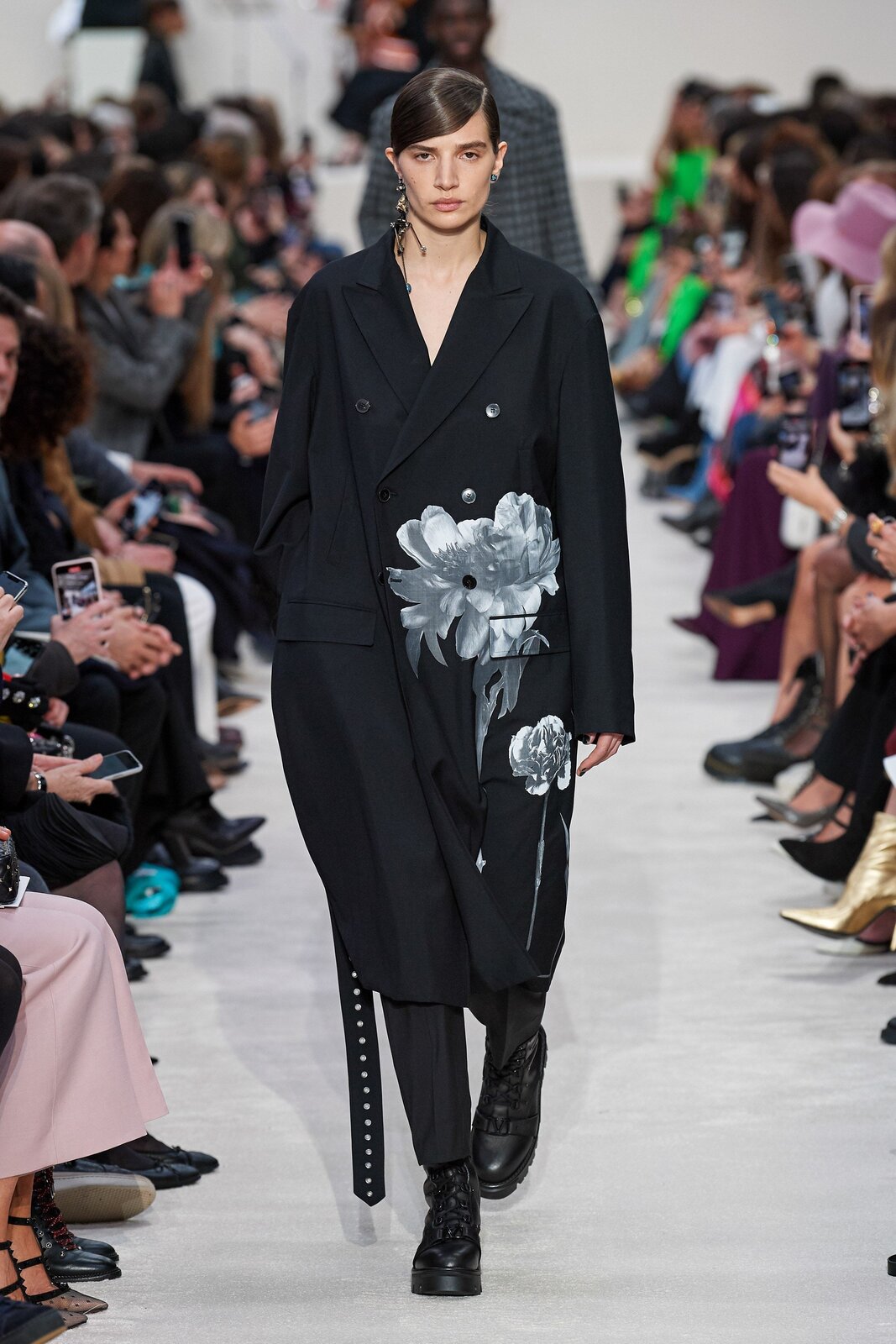 Fotografia przedstawia modelkę ubraną w luźny czarny płaszcz z dwoma jasnoszarymi kwiatowymi wzorami z jego lewej strony, czarne spodnie oraz czarne buty o grubej podeszwie. Dekolt płaszcza odsłania jasną skórę kobiety, a na jej uszach widoczne są kolczyki – na lewym uchu ozdoba jest mała i okrągła, na prawym zaś można dostrzec duży, wiszący kolczyk. Prawa dłoń modelki spoczywa w kieszeni płaszcza. Jej ciemne włosy rozdzielone są z boku przedziałkiem oraz zaczesane do tyłu. 