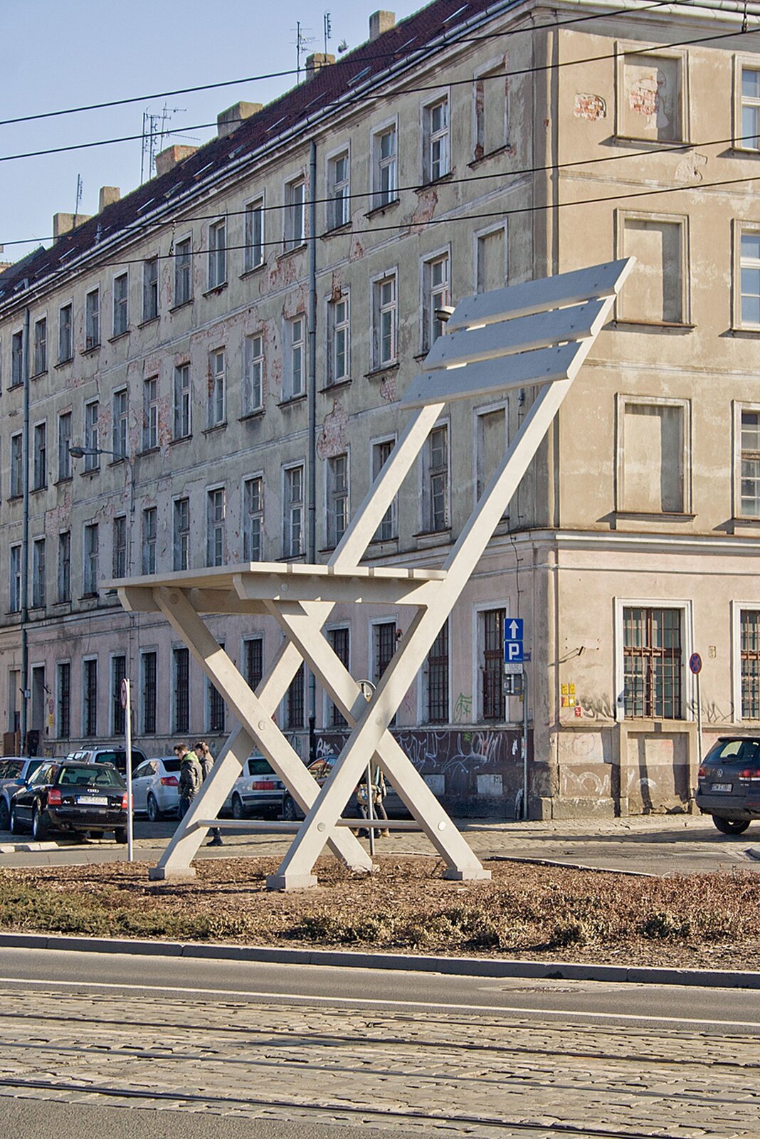 Ilustracja przedstawia krzesło powiększone do gigantycznych rozmiarów, stojące na ulicy. Jest to rzeźba plenerowa. Krzesło jest drewniane, białe, z oparciem, jest to krzesło składane. 