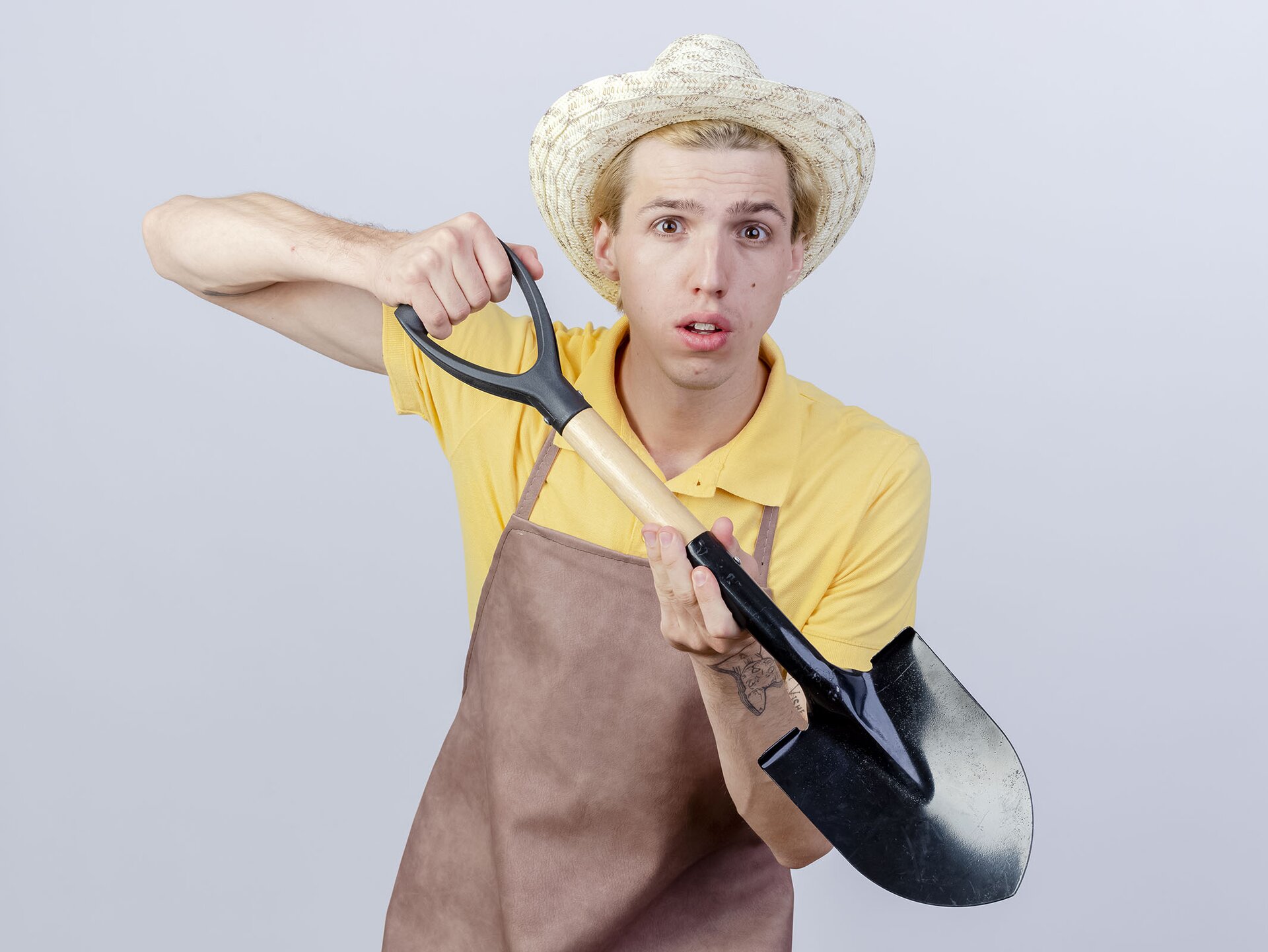 Zdjęcie przedstawia młodego mężczyznę w kapeluszu słomkowym i fartuchu, który trzyma w dłoniach łopatę. 
