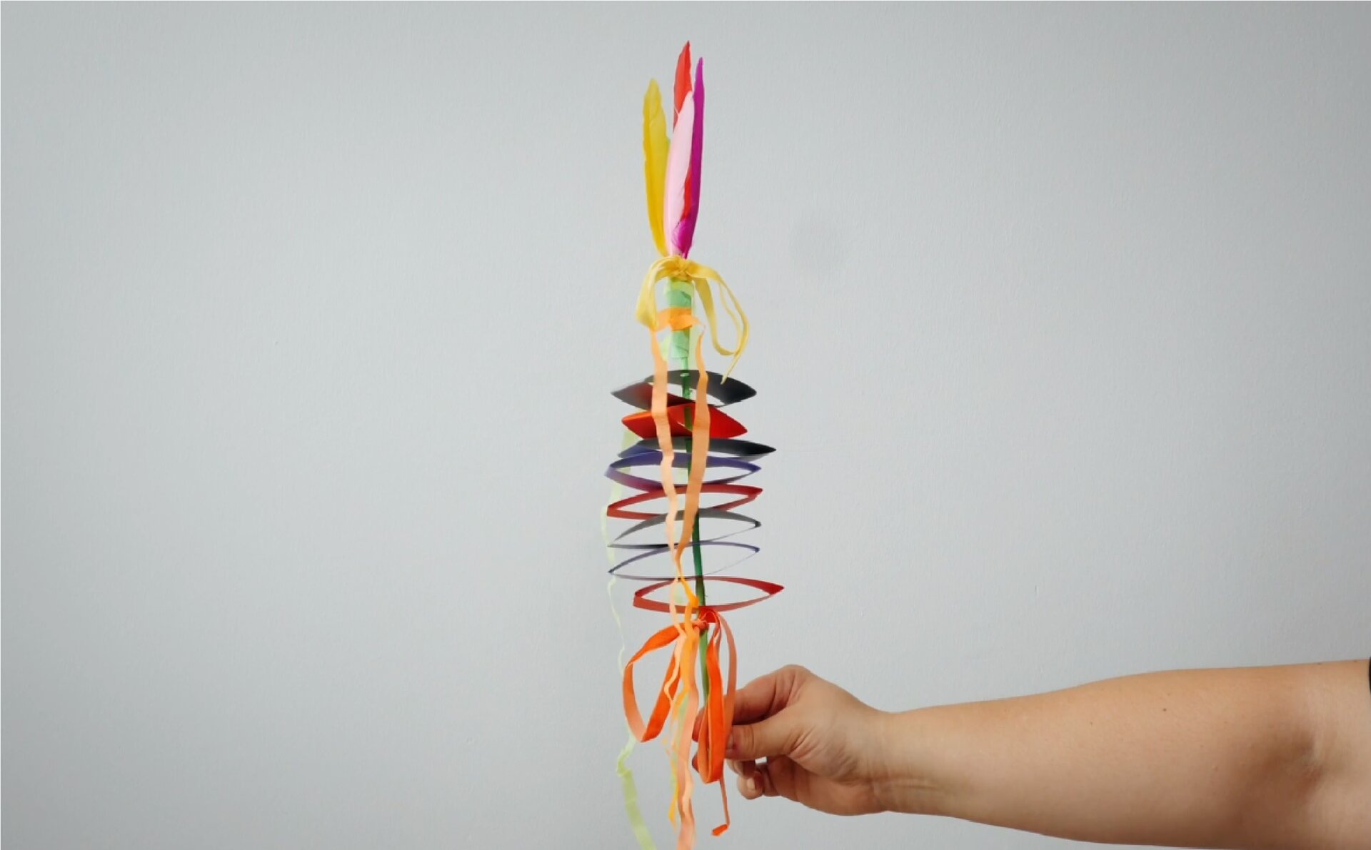 Zdjęcie przedstawia rękę trzymającą palmę wielkanocną. Tło jest jasnoszare. Palmę wykonano z kolorowych wstążek i kolorowego papieru. Jest taka sama, jak ta widoczna na filmie.