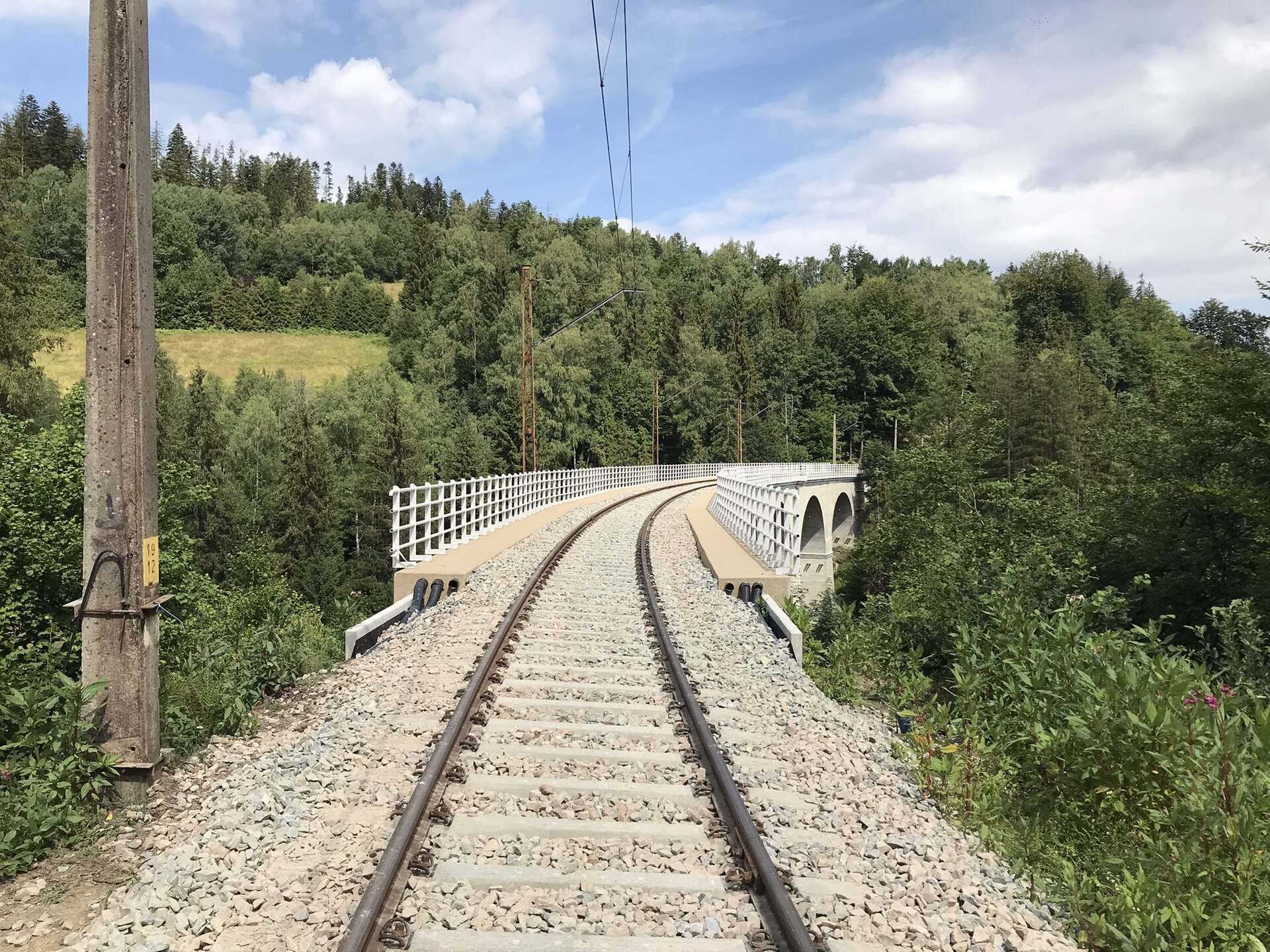 Zdjęcie przedstawia most łukowy. Most składa się z łuków połączonych z góry platformą. Na moście znajduje się barierka służąca do chronienia użytkowników mostu. Most posiada jeden tor kolejowy. Na moście znajdują się słupy elektryczne, które połączone są ze sobą liniami wysokiego napięcia. W tle widoczna jest zalesiona dolina oraz chmury na błękitnym niebie.