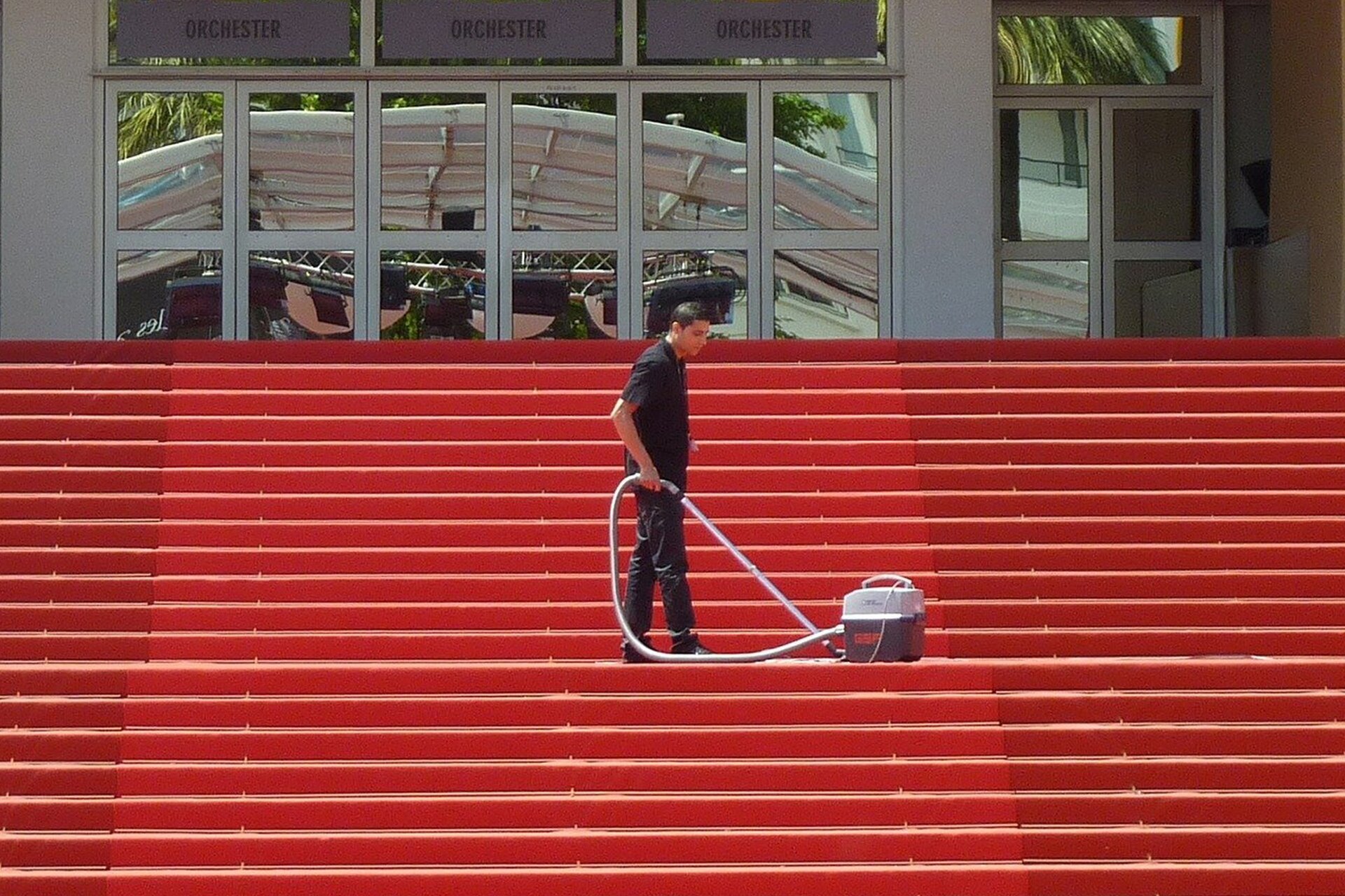 Na ilustracji obserwujmy młodego mężczyznę, który odkurza okryte czerwoną wykładziną schody prowadzące do budynku z dużymi szklanymi drzwiami. W odbiciu w tych drzwiach możemy dostrzec dach nad sceną.