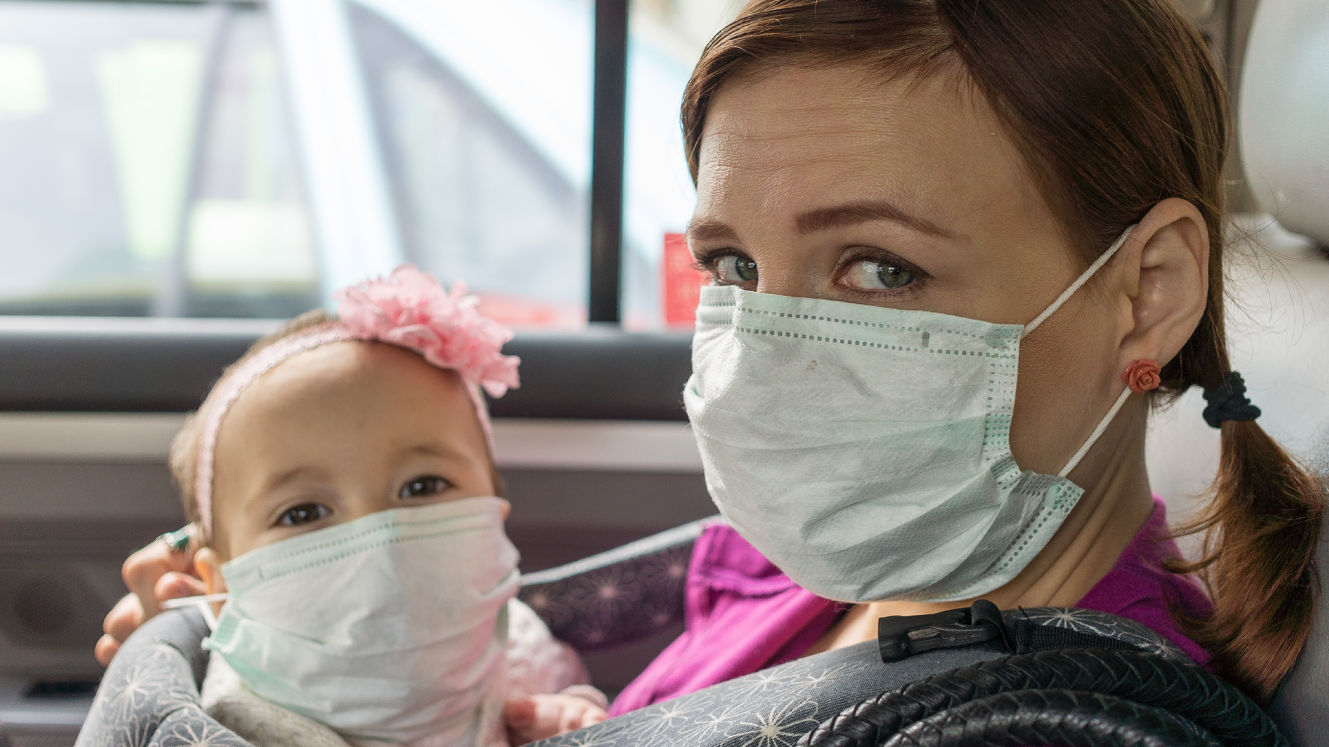 Zdjęcie przedstawia młodą kobietę oraz małą dziewczynkę, które siedzą w samochodzie. Kobieta i dziewczynka mają na twarzy maseczkę ochronną. Dziewczynka umieszczona jest w nosidełku, które kobieta ma na ramionach. Dziewczynka ma kokardę na głowie.