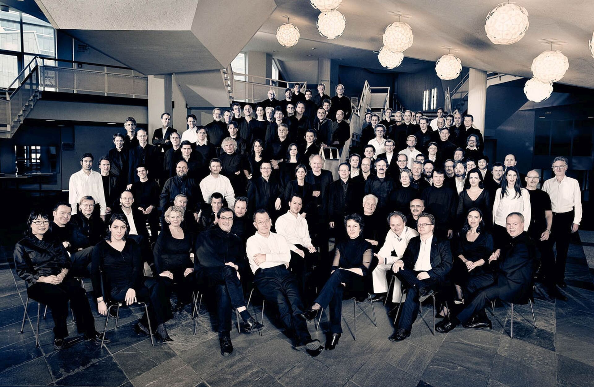 Ilustracja przedstawia orkiestrę Filharmonii Berlińskiej. Na zdjęciu widać kilkudziesięciu muzyków. Większość ubrana na czarno. Pojawiają się również osoby w białych koszulach.