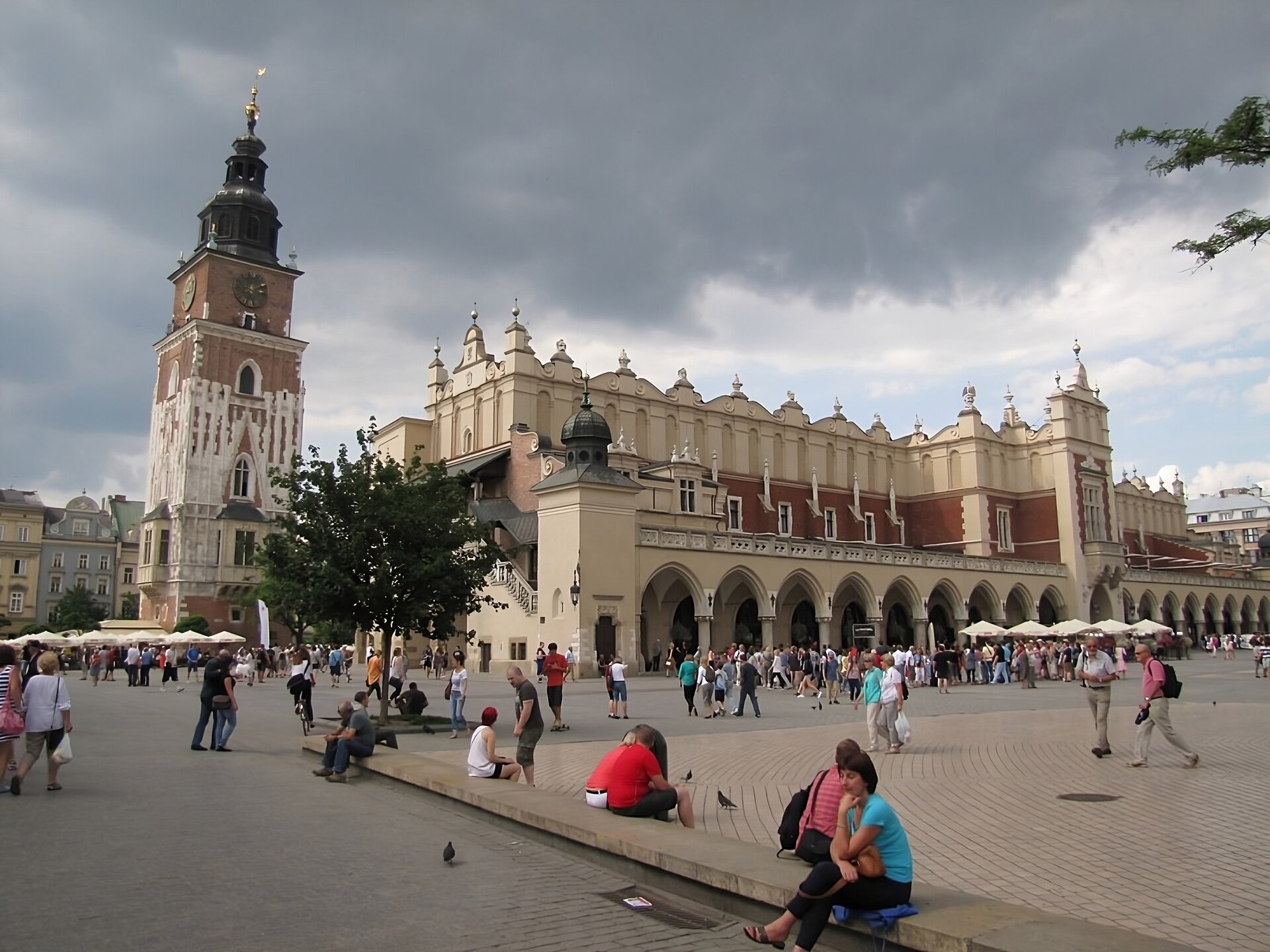 Zdjęcie przedstawia współczesny widok na krakowskie Sukiennice i wieżę dawnego ratusza miejskiego. Renesansowa zabudowa sukiennic i widocznej jej ściany frontowej od strony Kościoła Mariackiego wyróżnia się arkadami na parterze o pełnych łukach otworów i bogato zdobioną attyką u szczytu. Pomiędzy arkadami a attyką długa wnęka ściany z otworami okiennymi i wykusz pośrodku z centralnym wejściem do wnętrza obiektu. Skrzydło wschodnie częściowo zasłonięte przez drzewo na rynku, widać jednak jedną z wież narożnych sukiennic z hełmem na szczycie. Bliżej lewej krawędzi fotografii wieża ratuszowa ceglana, uzupełniona kamieniem w połowie wysokości. Wieża zwęża się ku szczytowi. Poniżej kopuły z hełmem widoczne tarcze zegarowe na dwóch ścianach pod szczytem. U dołu wieży ratuszowej wykusze z oknami. Na pierwszym planie fotografii widać grupy przechodniów i siedzących na ławach turystów. 