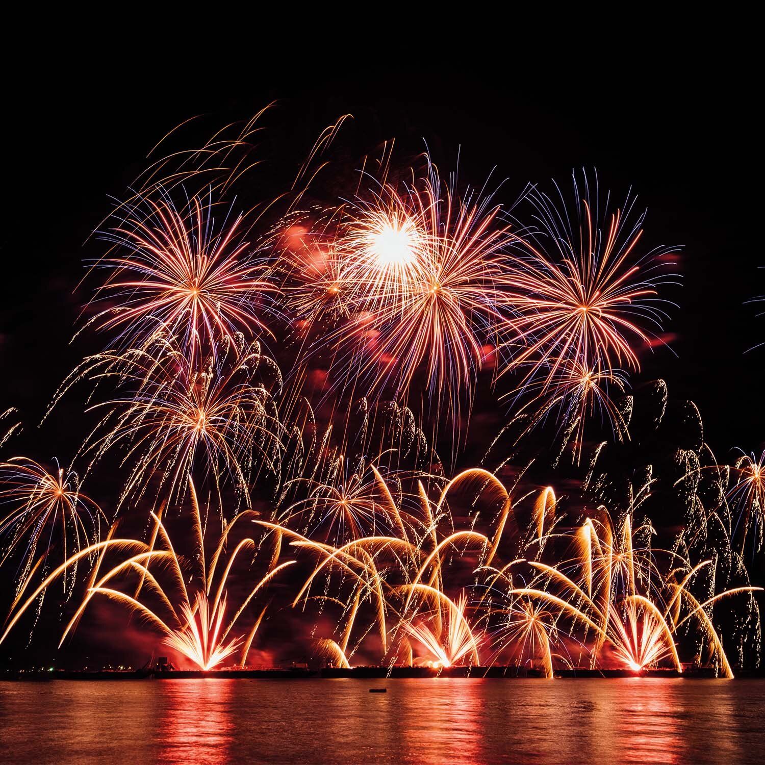 Zdjęcie przedstawia nocny pokaz fajerwerków nad brzegiem zbiornika wodnego. Na pierwszym planie widoczna jest tafla wody, w której odbijają się sztuczne ognie. Na dalszym planie widoczne są kolorowe fajerwerki na dwóch poziomach, tuż nad taflą wody i na niebie.