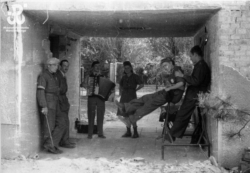 Fotografia przedstawiająca kilkoro ludzi stojących w bramie. Kobieta w środku, której towarzyszy pięciu mężczyzn, dwóch po jej lewej, gdzie jeden siedzi na metalowej ramie, drugi zaś stoi za nim; po jej prawej trzech pozostałych, najbliższy z akordeonem, dalej dwoje podpiera ścianę, ostatni trzyma laskę.