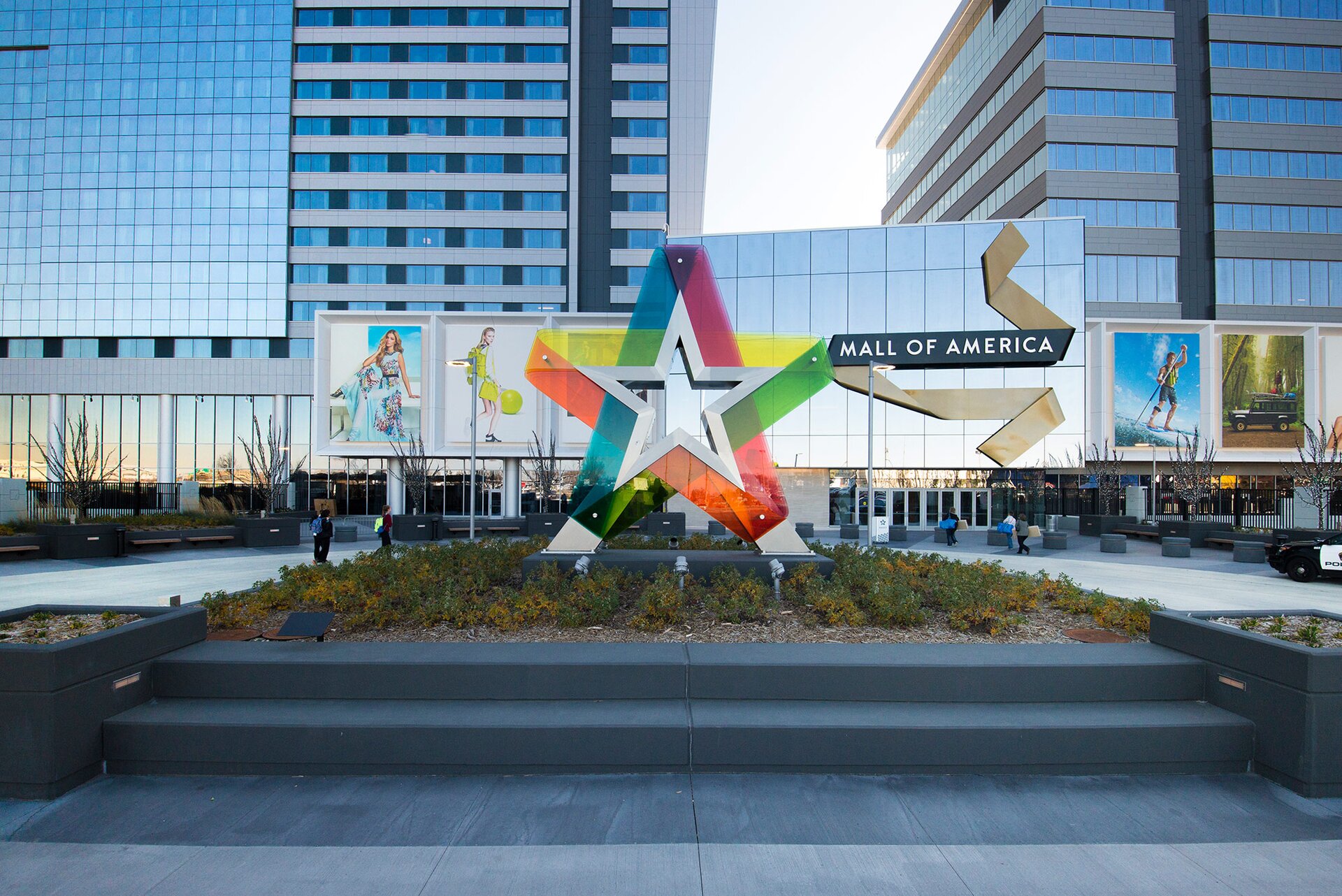 Zdjęcie przedstawia nowoczesny, przeszklony budynek składający się z niskiej części oraz dwóch wielopiętrowych wieżowców po bokach. Pośrodku nad wejściami w niskiej części budynku widoczna jest prostokątna elewacja ze szkła, na której znajduje się napis w języku angielskim: MALL OF AMERICA. Przed wejściem widoczne są ogromne klomby. W centralnej części największego klombu widoczna jest rzeźba w kształcie pięcioramiennej srebrnej gwiazdy otoczonej różnokolorowymi, półprzeźroczystymi pasami. 