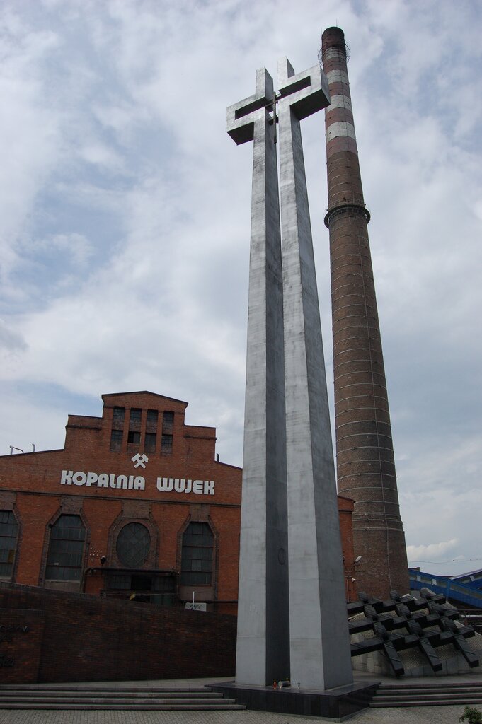 Zdjęcie przedstawia pomnik - wysoki, betonowy krzyż. Za nim znajduje się budynek z czerwonej cegły z napisem Kopalnia Wujek.