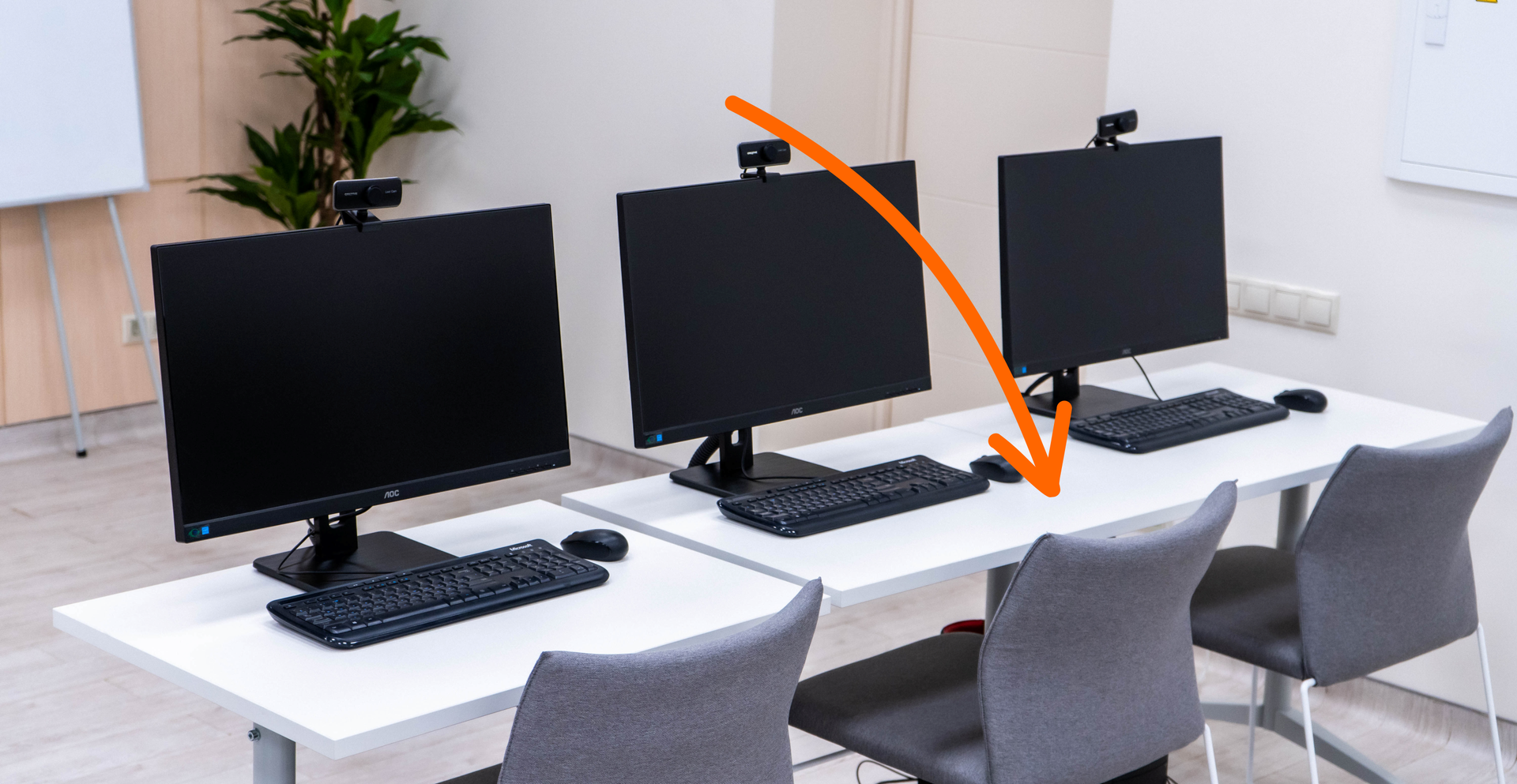 Zdjęcie przedstawia trzy biurka obok siebie, na których znajdują się monitory i klawiatury komputera. Przy każdym biurku stoi krzesło. Strzałka na ilustracji skierowana jest na biurko.