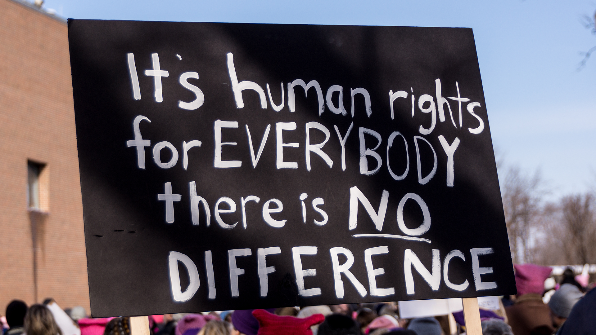 Zdjęcie stojącego tłumu ludzi zakrywa napis wykonany na czarnym tle białymi literami. Napis w języku angielskim głosi: IT'S HUMAN RIGHTS FOR EVERYBODY THERE IS NO DIFFERENCE. 